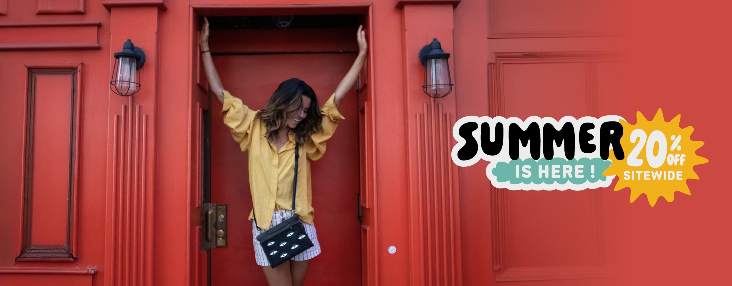 Model standing in front of a red building wearing a Min & Mon crossbody bag in the Summer is Here campaign featuring 20% OFF sitewide.