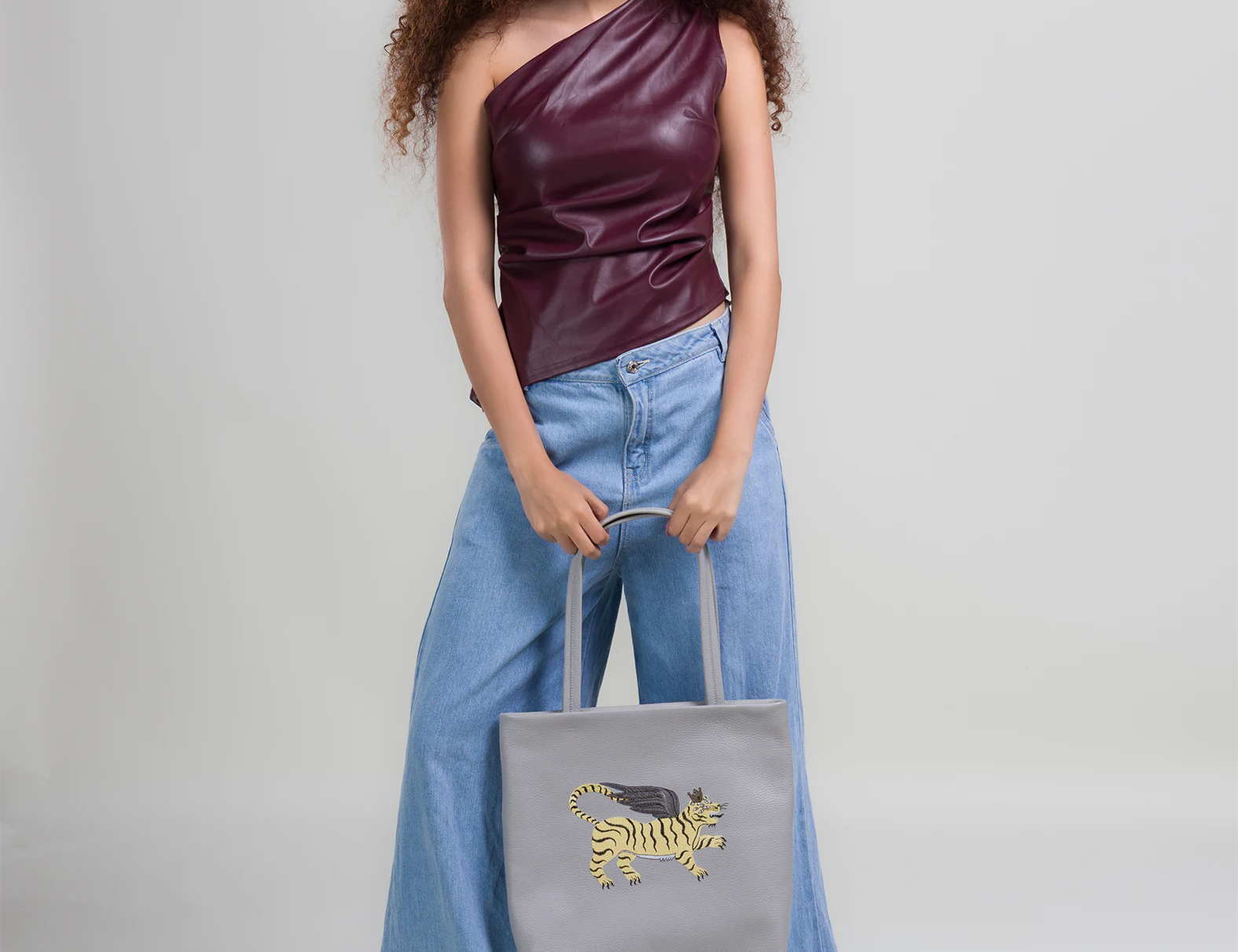 Woman wearing a one-shoulder top and wide-leg jeans, holding a gray tote bag with a tiger design.