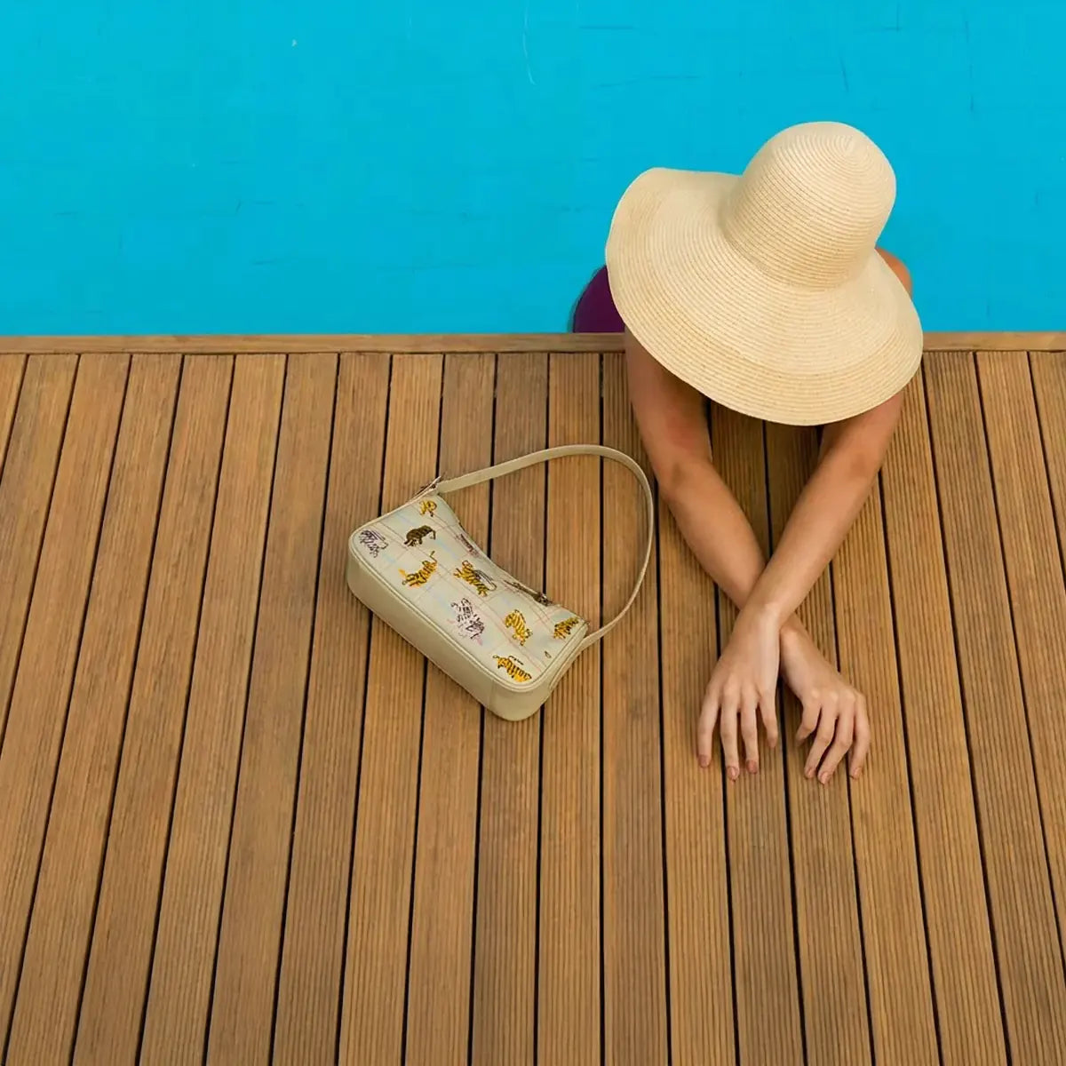 Overhead poolside photo featuring a woman in a sunhat beside a printed Min & Mon shoulder bag on a wooden deck.