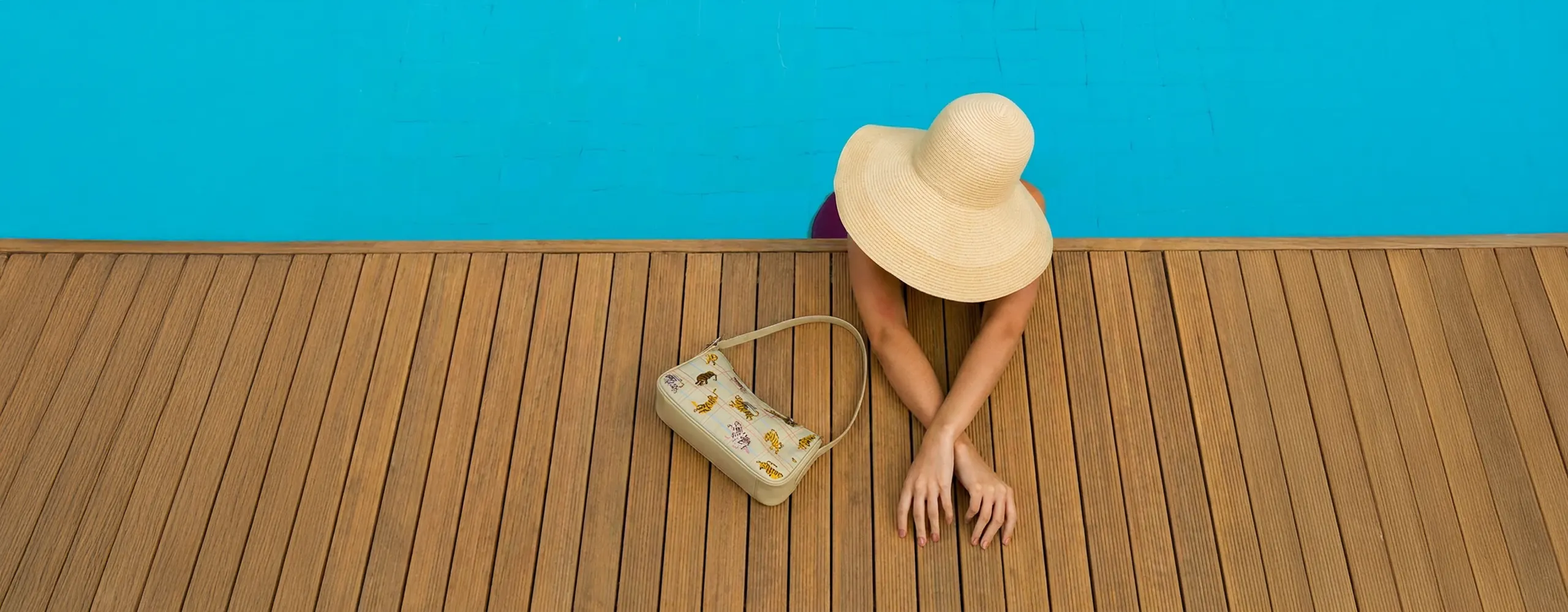 Overhead poolside photo featuring a woman in a sunhat beside a printed Min & Mon shoulder bag on a wooden deck.