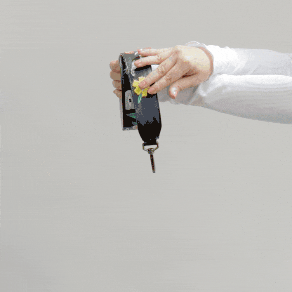 Hand holds and rotates a black embroidered bag strap with floral and eye motifs against a neutral background, showing texture and hardware details in motion.