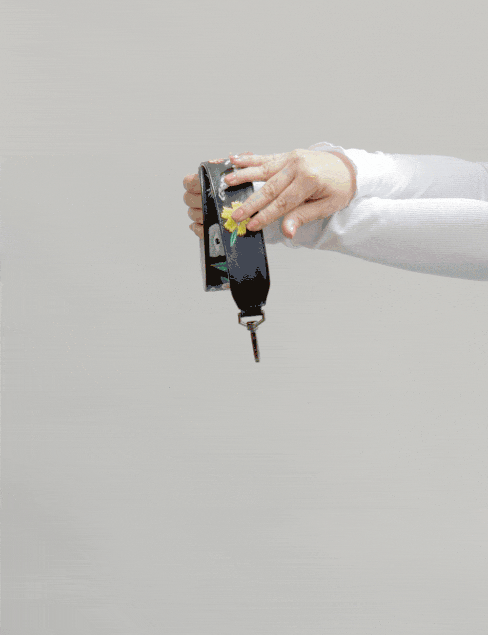 Hand holds and rotates a black embroidered bag strap with floral and eye motifs against a neutral background, showing texture and hardware details in motion.