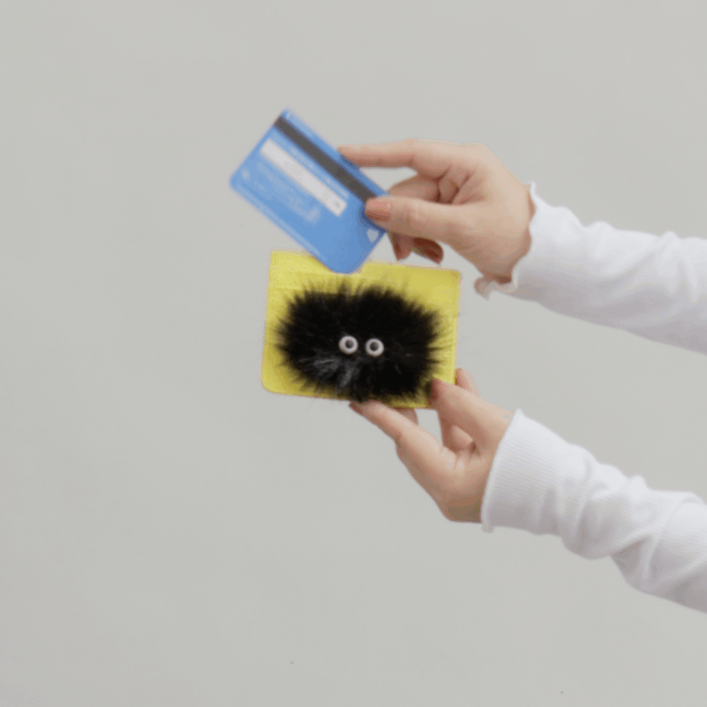 Hands hold a yellow card holder with a playful black fuzzy creature design while inserting a blue credit card, demonstrating card access against a neutral background.
