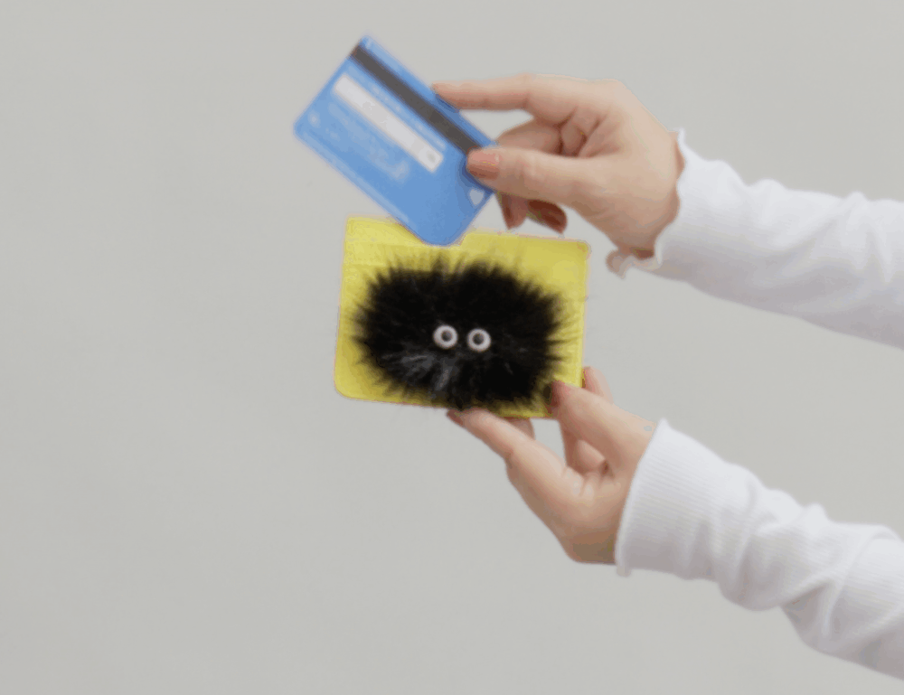 Hands hold a yellow card holder with a playful black fuzzy creature design while inserting a blue credit card, demonstrating card access against a neutral background.