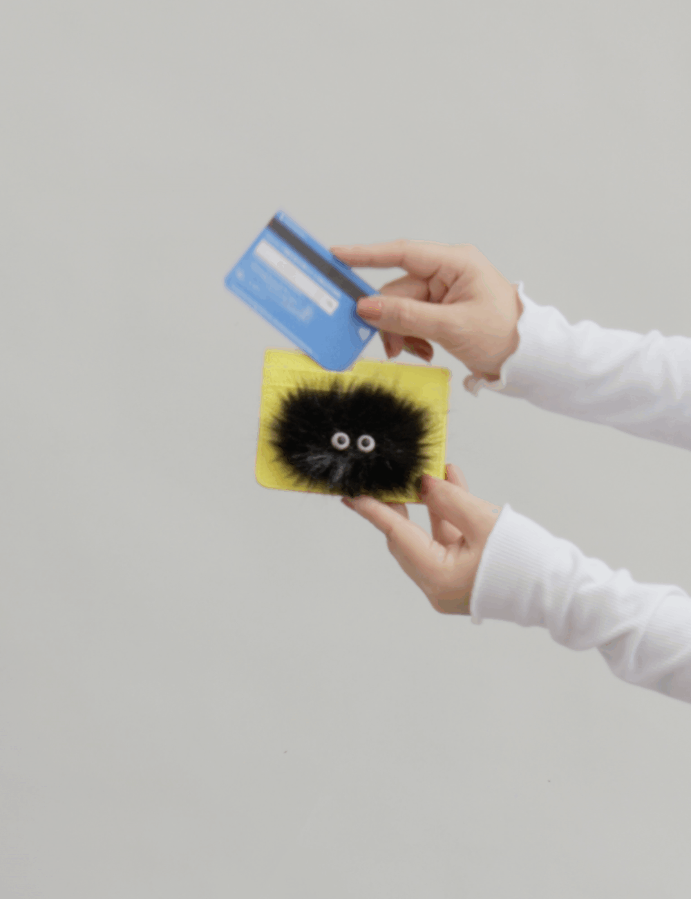 Hands hold a yellow card holder with a playful black fuzzy creature design while inserting a blue credit card, demonstrating card access against a neutral background.