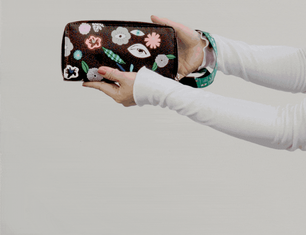 Hands hold and gently tilt a black zip-around wallet with colorful embroidered flowers and eye motifs, showcasing the design and wrist strap against a neutral background.
