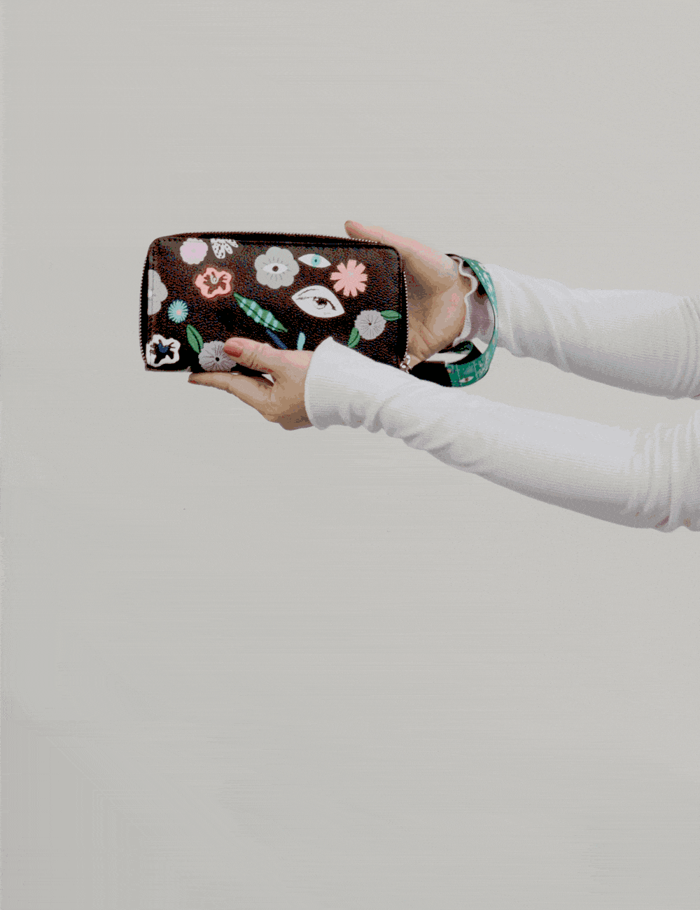 Hands hold and gently tilt a black zip-around wallet with colorful embroidered flowers and eye motifs, showcasing the design and wrist strap against a neutral background.