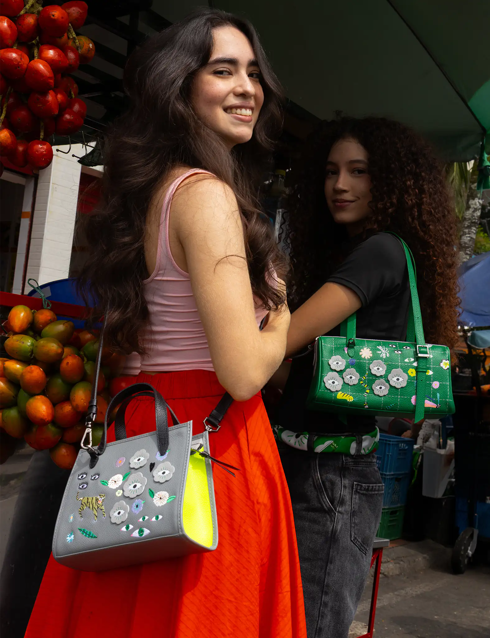 Two women with long hair stand smiling, each carrying colorful handbags with floral designs. They are near a fruit stand with stacks of mangos. The scene is vibrant and lively.