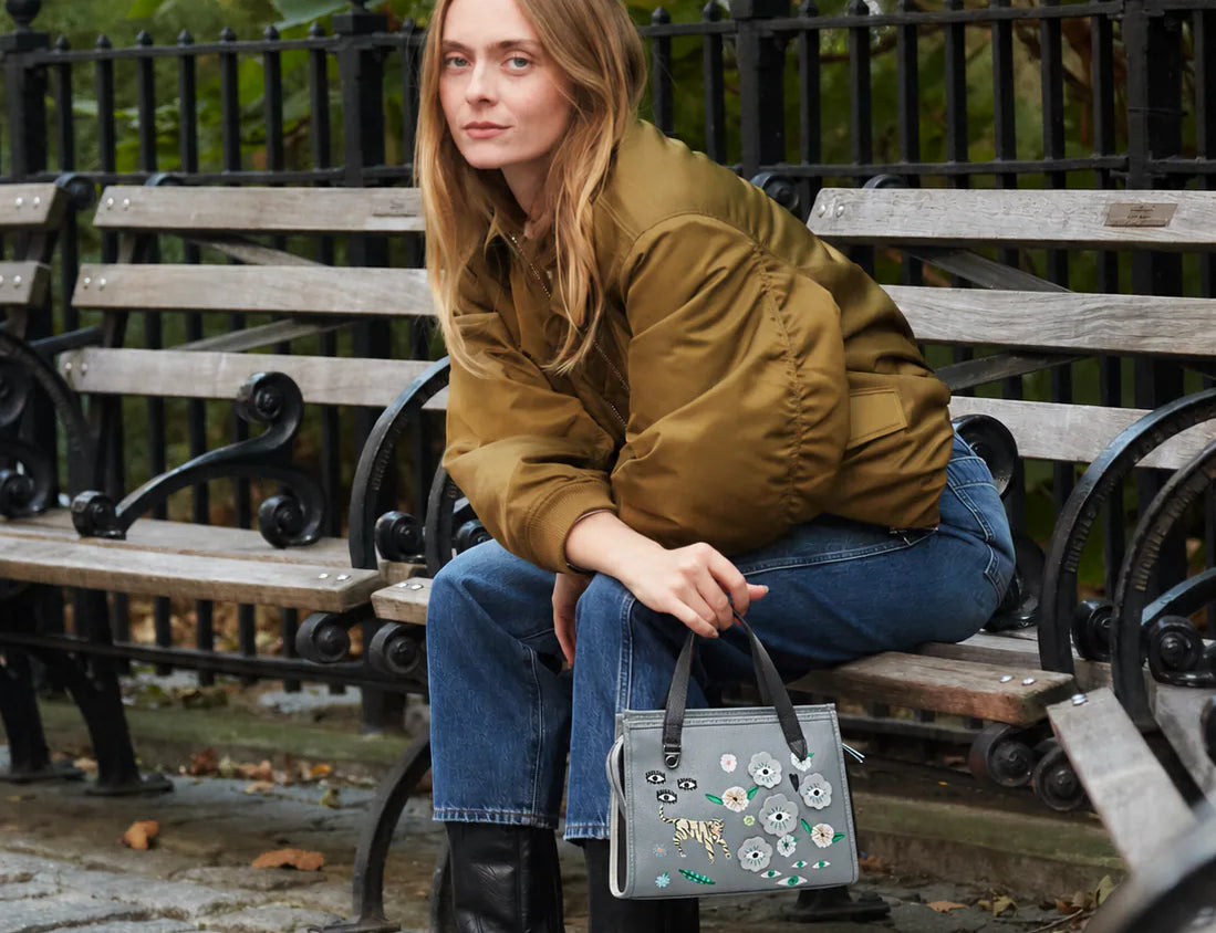 Woman sitting on a bench in an urban park holding a gray handbag with floral design.