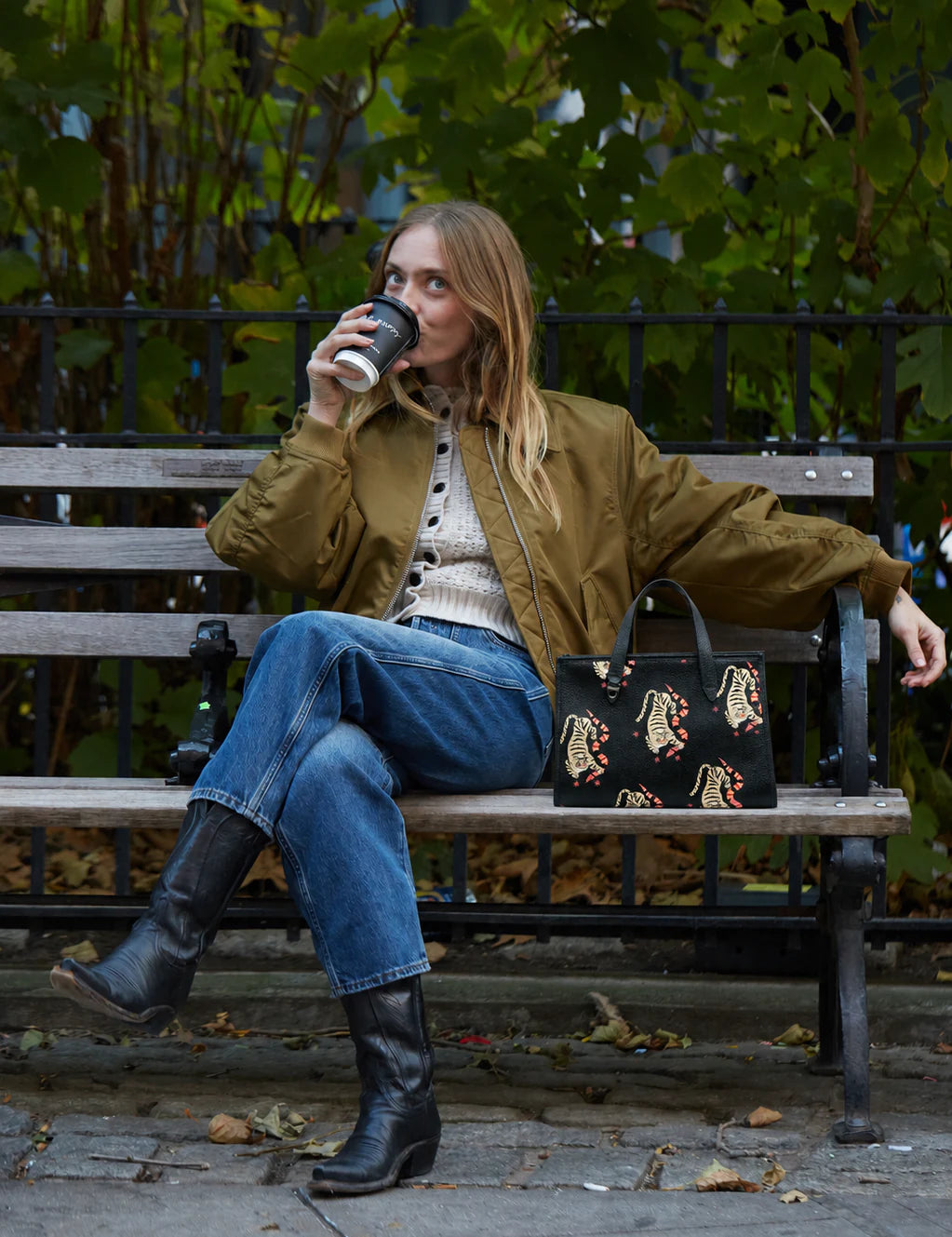 Woman in an olive bomber jacket sits on a park bench sipping coffee, with a black tiger-print bag beside her; green foliage and a black iron fence fill the background.