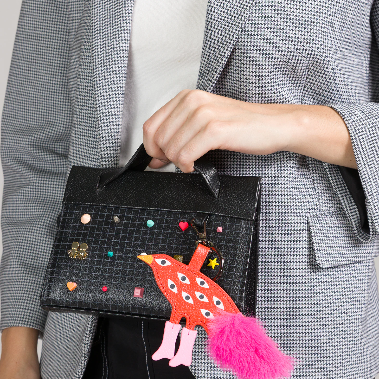 Close-up of a black top-handle handbag with a grid pattern, colorful studs, and a playful red bird charm with multiple eyes and a pink tail, held by a person in a gray blazer.
