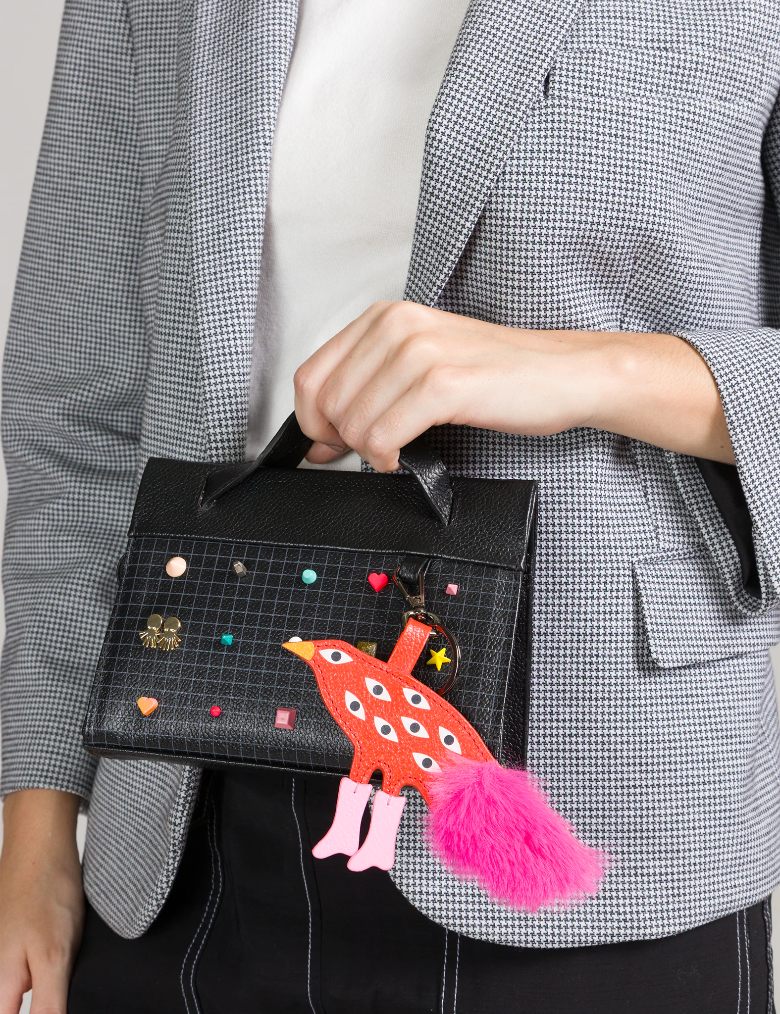 Close-up of a black top-handle handbag with a grid pattern, colorful studs, and a playful red bird charm with multiple eyes and a pink tail, held by a person in a gray blazer.