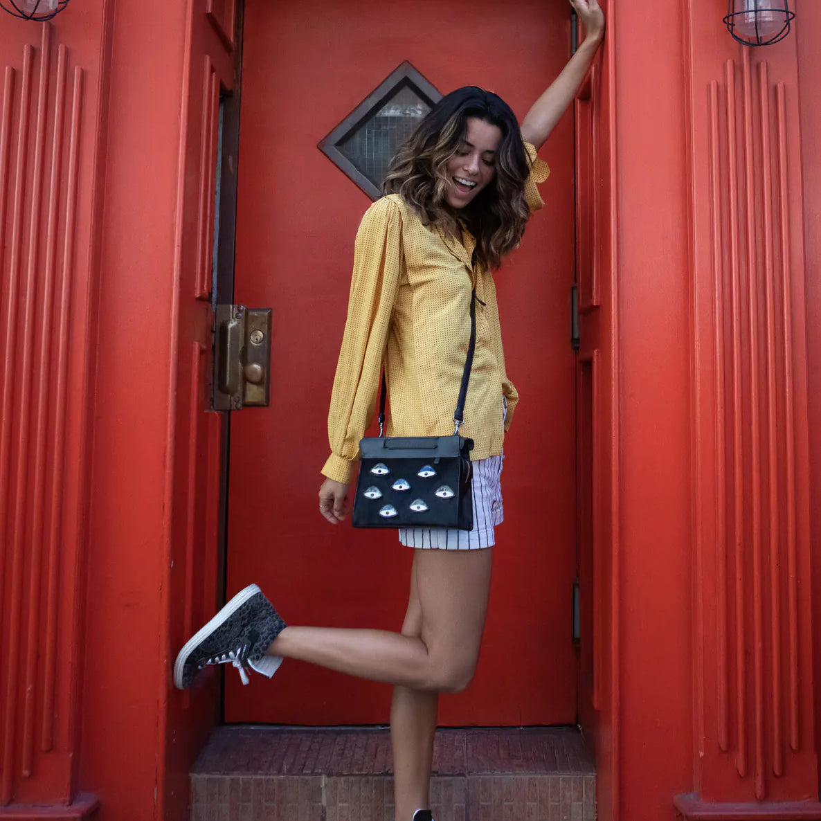 Woman in a yellow blouse and striped shorts poses in a doorway with a bright red frame, wearing a black crossbody bag with eye motifs and lifting one leg playfully.