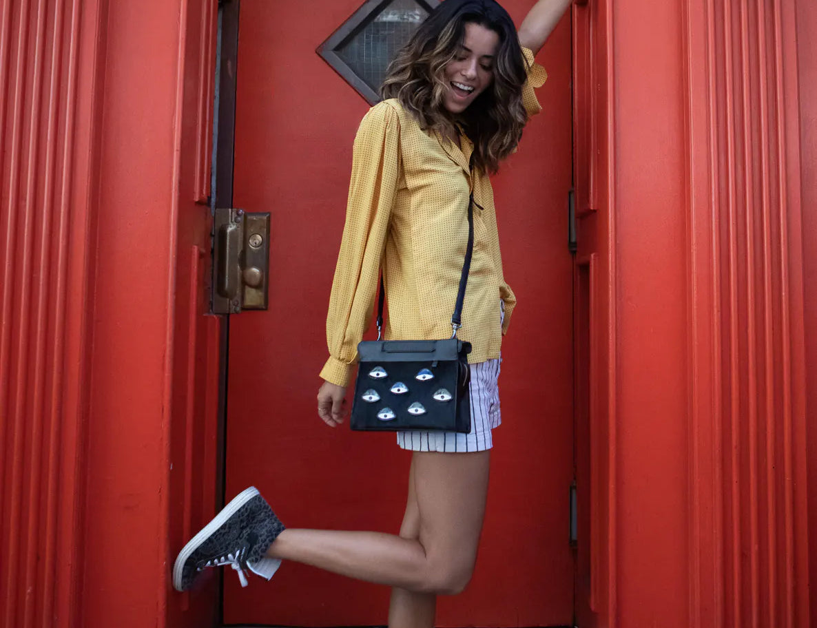 Woman in a yellow blouse and striped shorts poses in a doorway with a bright red frame, wearing a black crossbody bag with eye motifs and lifting one leg playfully.