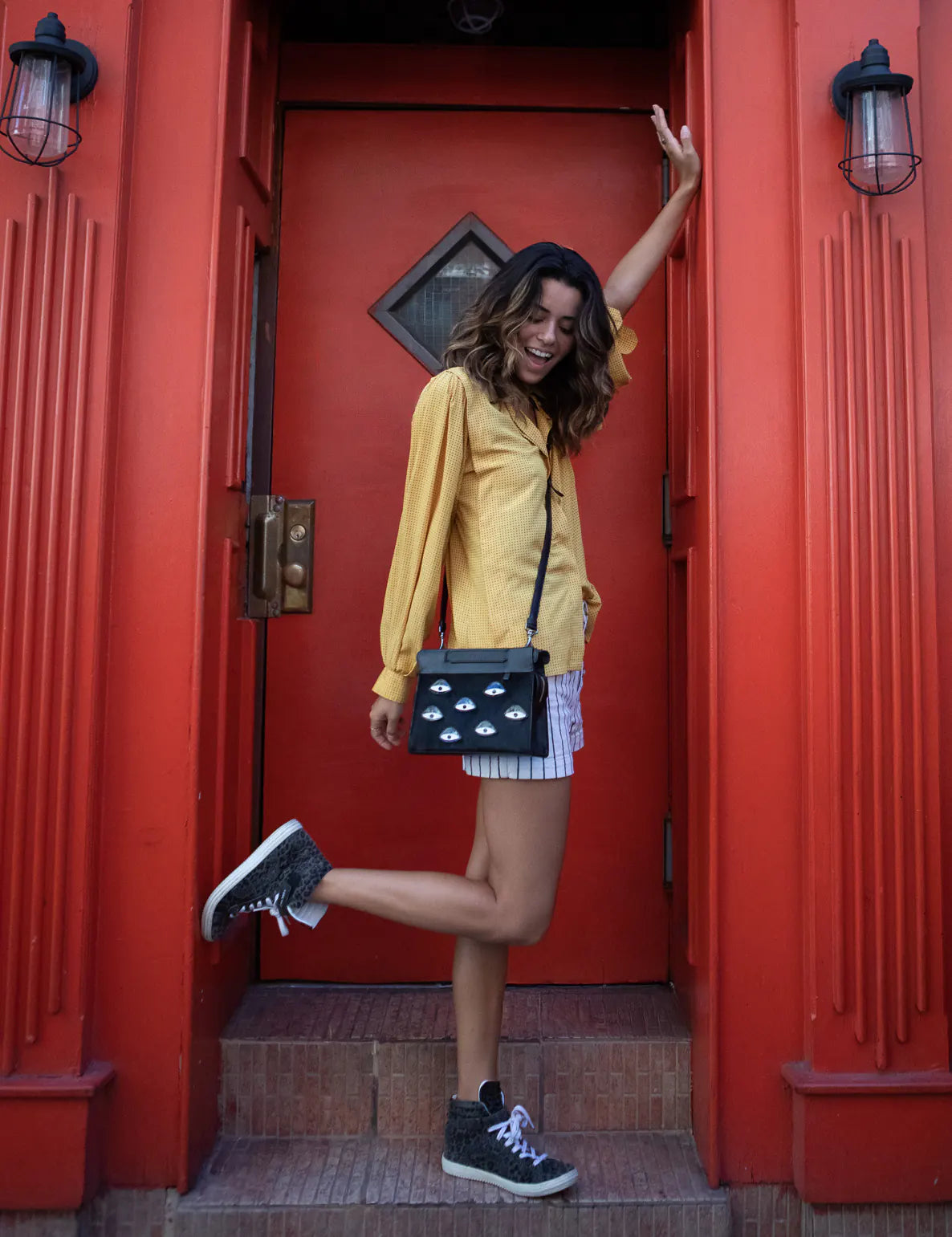 Woman in a yellow blouse and striped shorts poses in a doorway with a bright red frame, wearing a black crossbody bag with eye motifs and lifting one leg playfully.