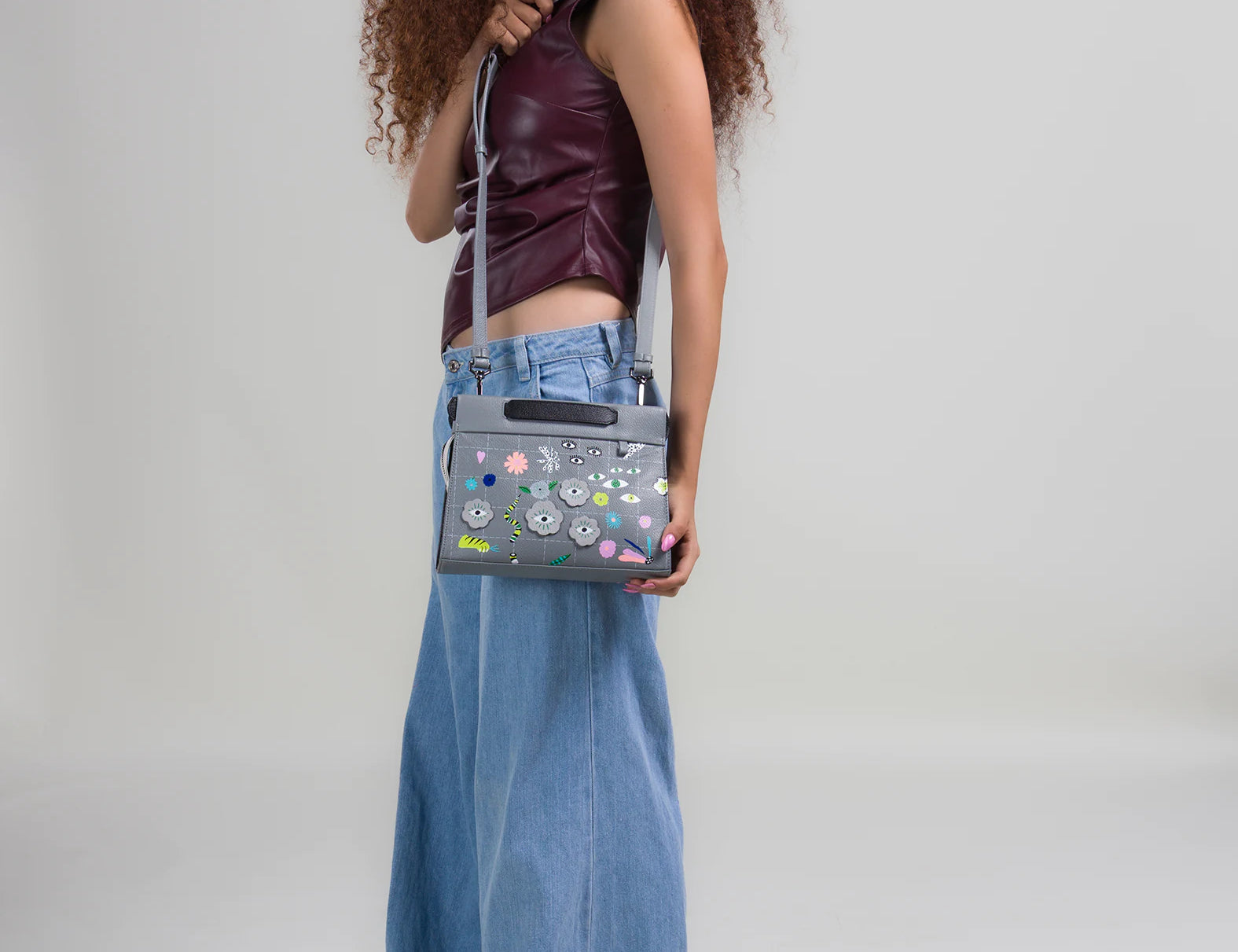 Woman with curly hair in a burgundy sleeveless top and wide-leg jeans looks down at a gray handbag with colorful floral embroidery, worn crossbody against a neutral background.