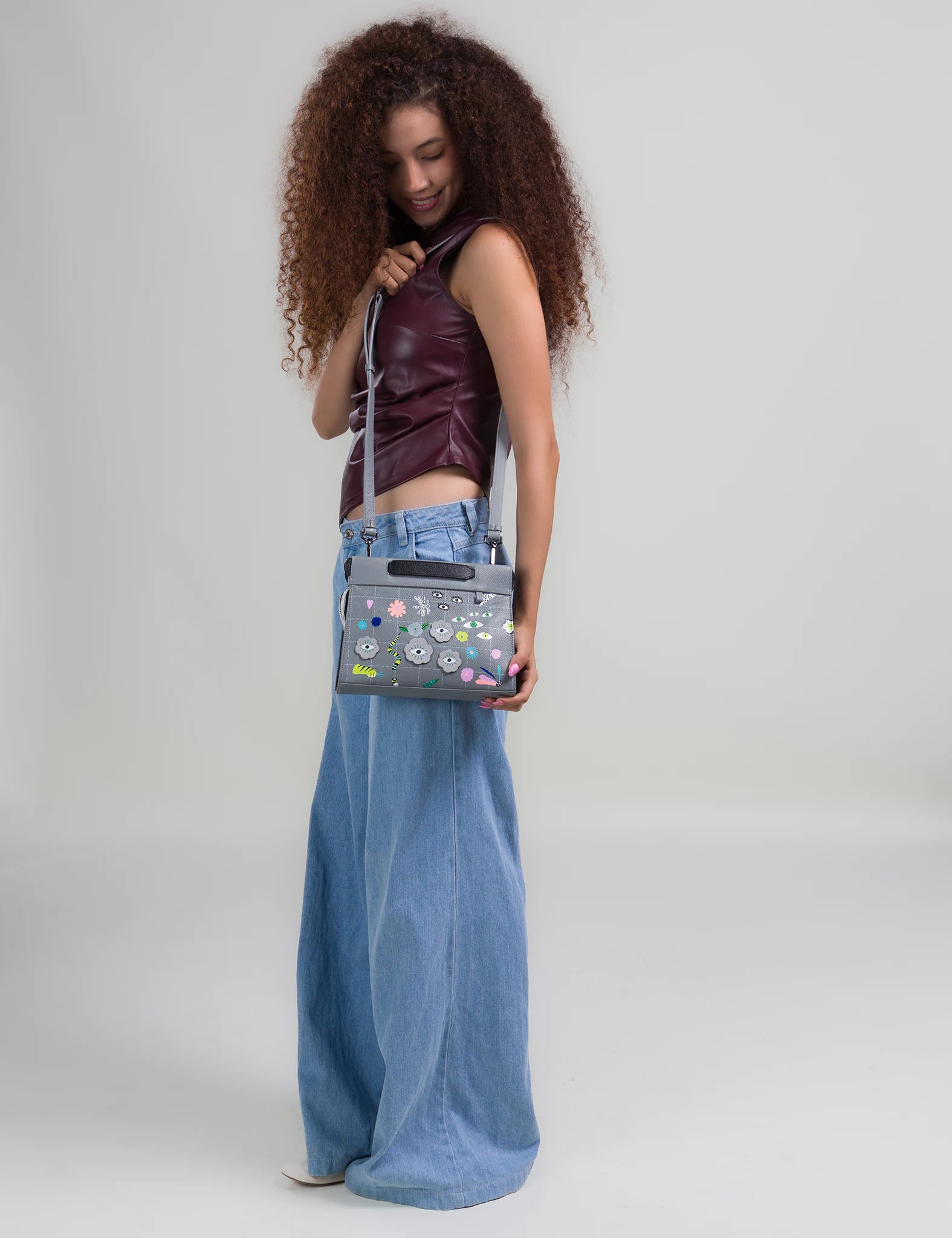 Woman with curly hair in a burgundy sleeveless top and wide-leg jeans looks down at a gray handbag with colorful floral embroidery, worn crossbody against a neutral background.