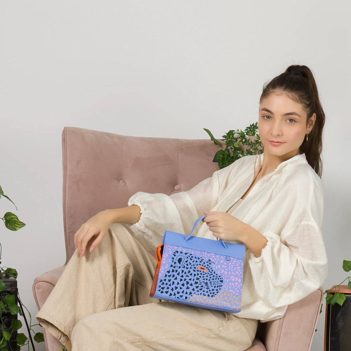 Model sitting in a pink armchair holding the Min and Mon Vali handbag featuring a laser-cut jaguar design for the Wild Heart collection. Retro set with vintage televisions and climbing plants.