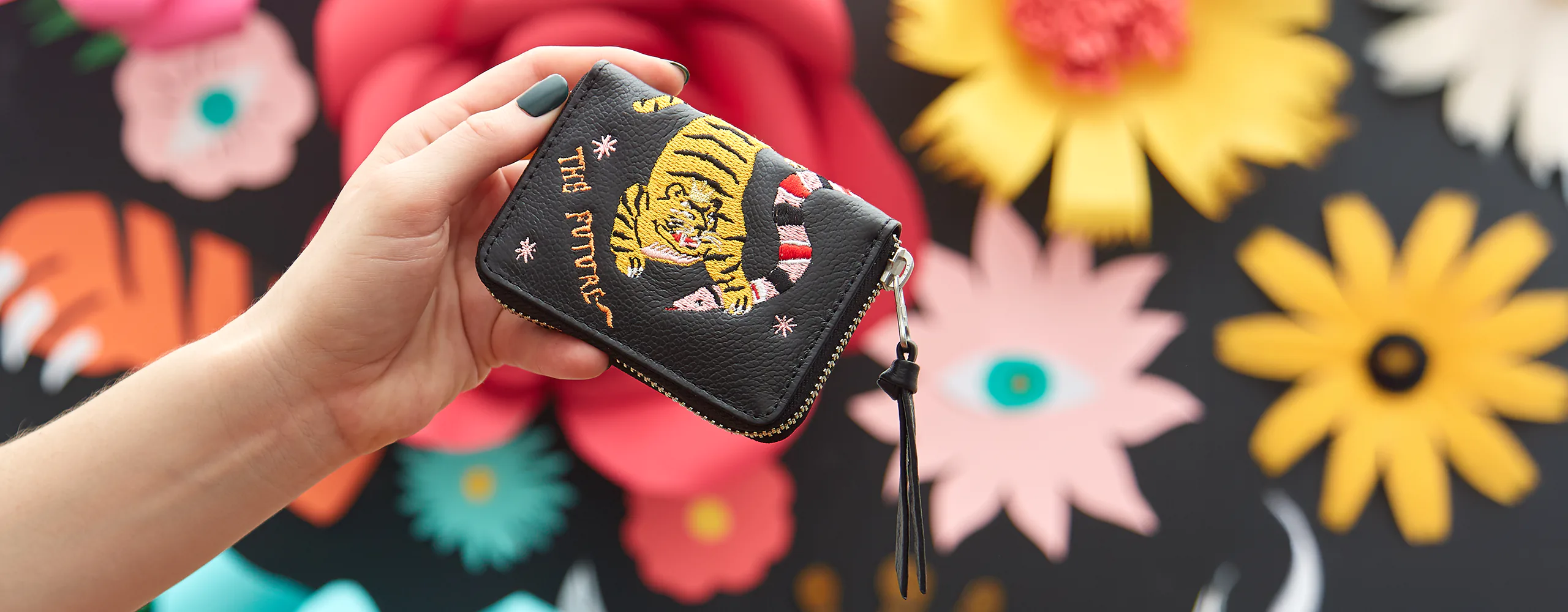Close-up of a Min & Mon zip wallet with tiger embroidery held in front of a colorful floral mural.