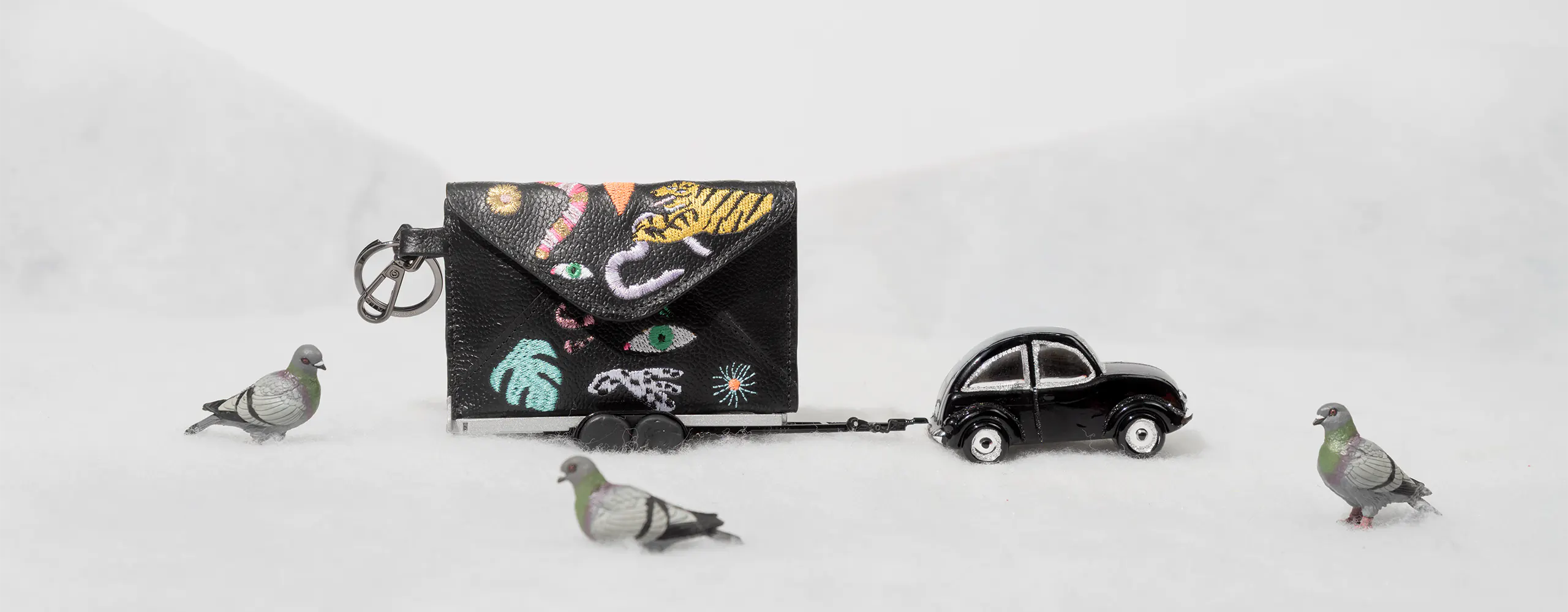 Black leather wallet with embroidered playful icons, styled like a tiny trailer pulled by a toy car, surrounded by pigeons on a snowy surface.