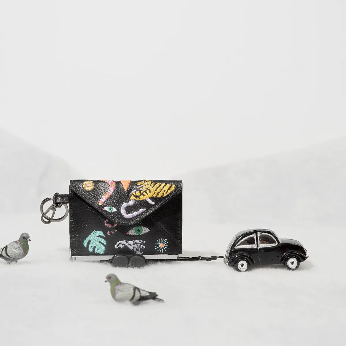 Mini black leather wallet with colorful embroidered motifs, styled in a whimsical winter scene with pigeons and a toy car.