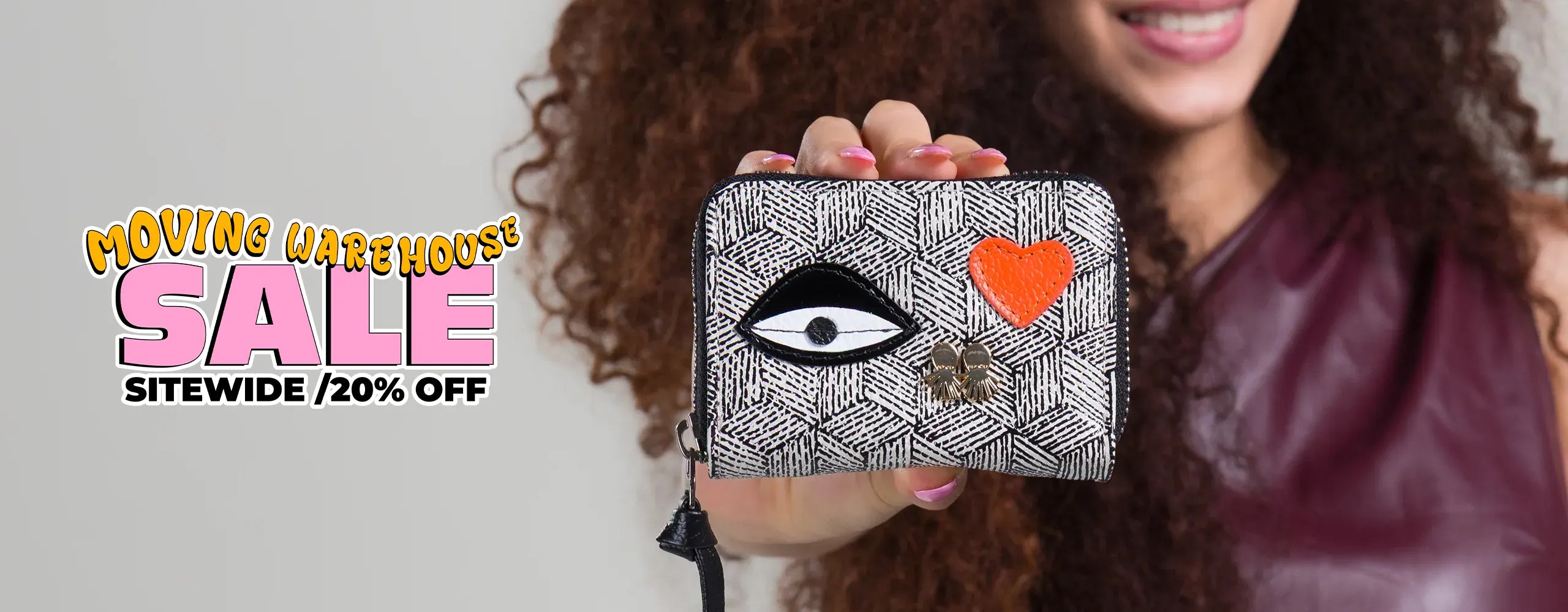 Moving Warehouse Sale horizontal banner for the wallet collection with 20% off sitewide. A model smiles while showing a close-up of a compact monochrome geometric pattern wallet with an eye patch and an orange heart.