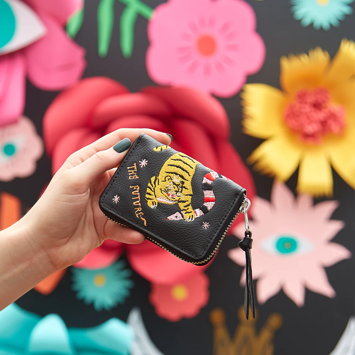 Close-up of a Min & Mon zip wallet with tiger embroidery held in front of a colorful floral mural.