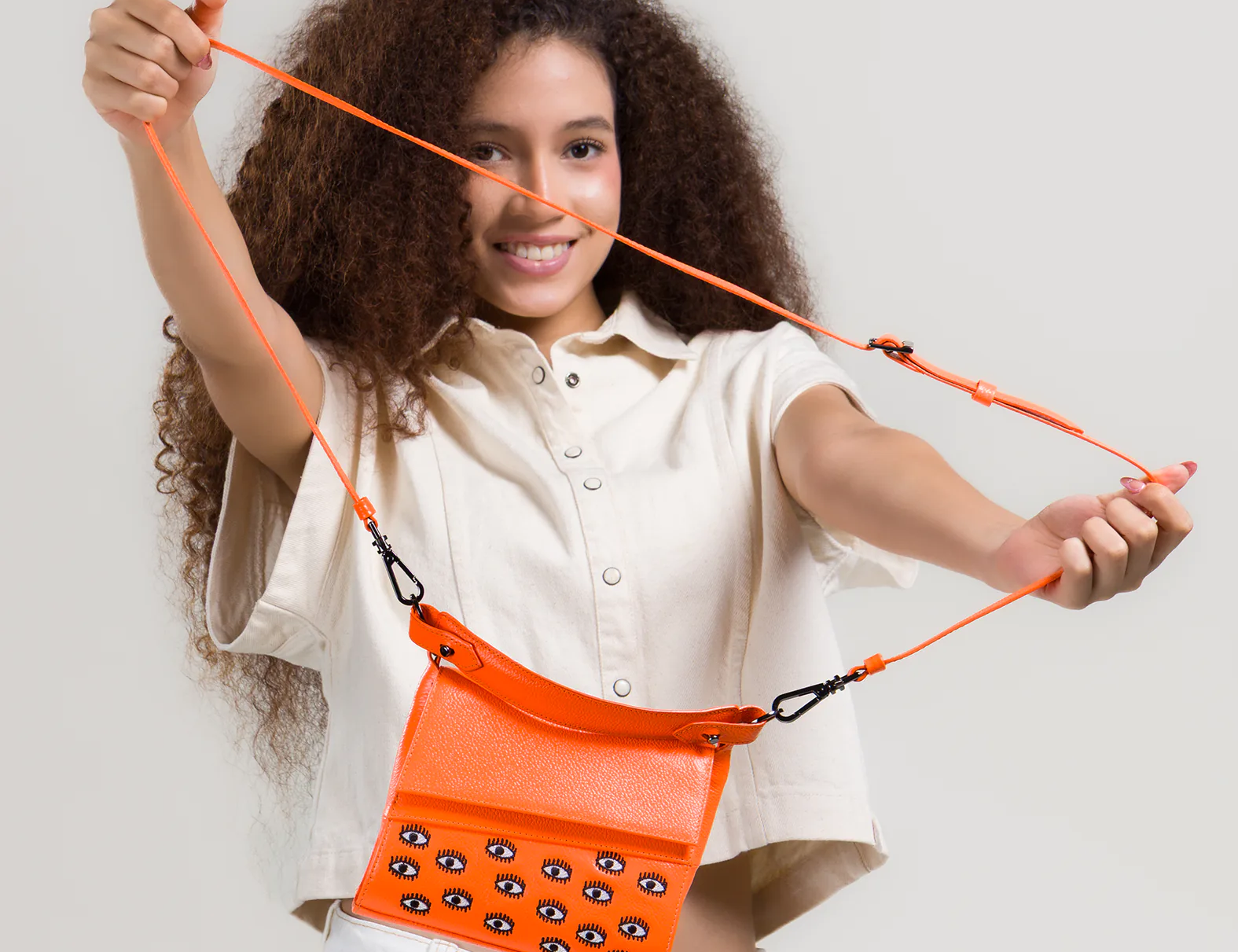 Smiling woman with curly hair stretches the strap of a bright orange crossbody bag featuring small embroidered eye motifs, wearing a light short-sleeve top and white jeans.