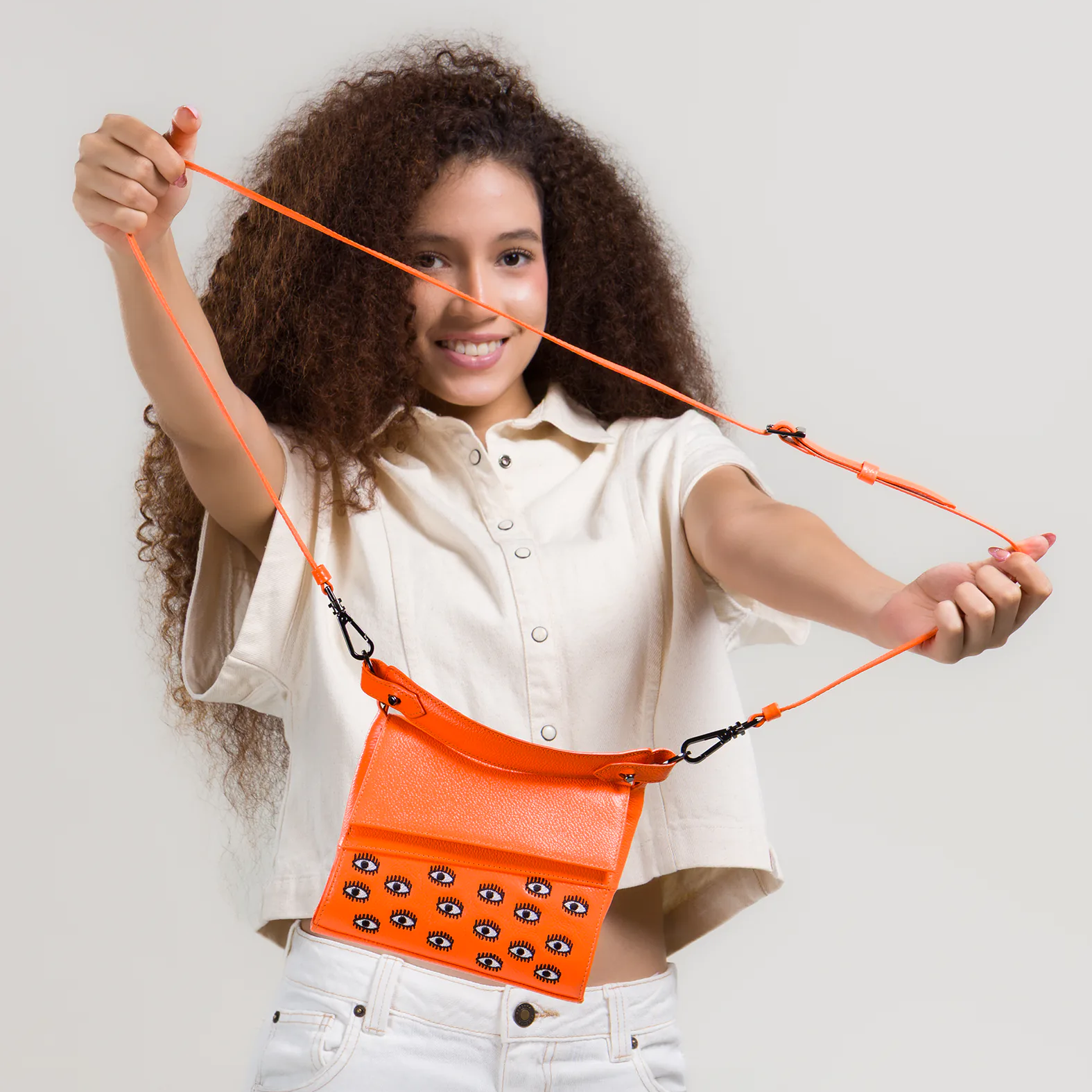 Smiling woman with curly hair stretches the strap of a bright orange crossbody bag featuring small embroidered eye motifs, wearing a light short-sleeve top and white jeans.