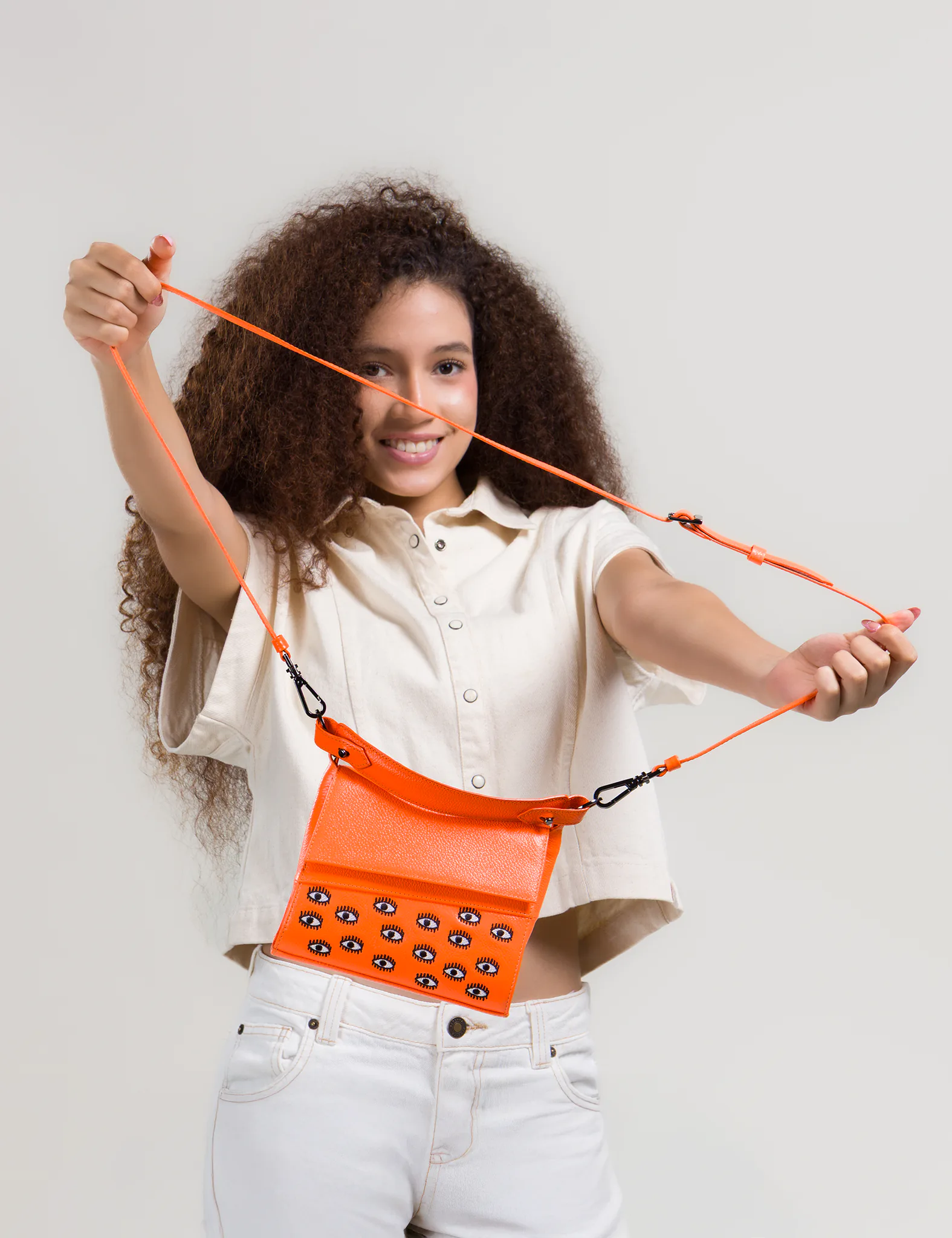 Smiling woman with curly hair stretches the strap of a bright orange crossbody bag featuring small embroidered eye motifs, wearing a light short-sleeve top and white jeans.