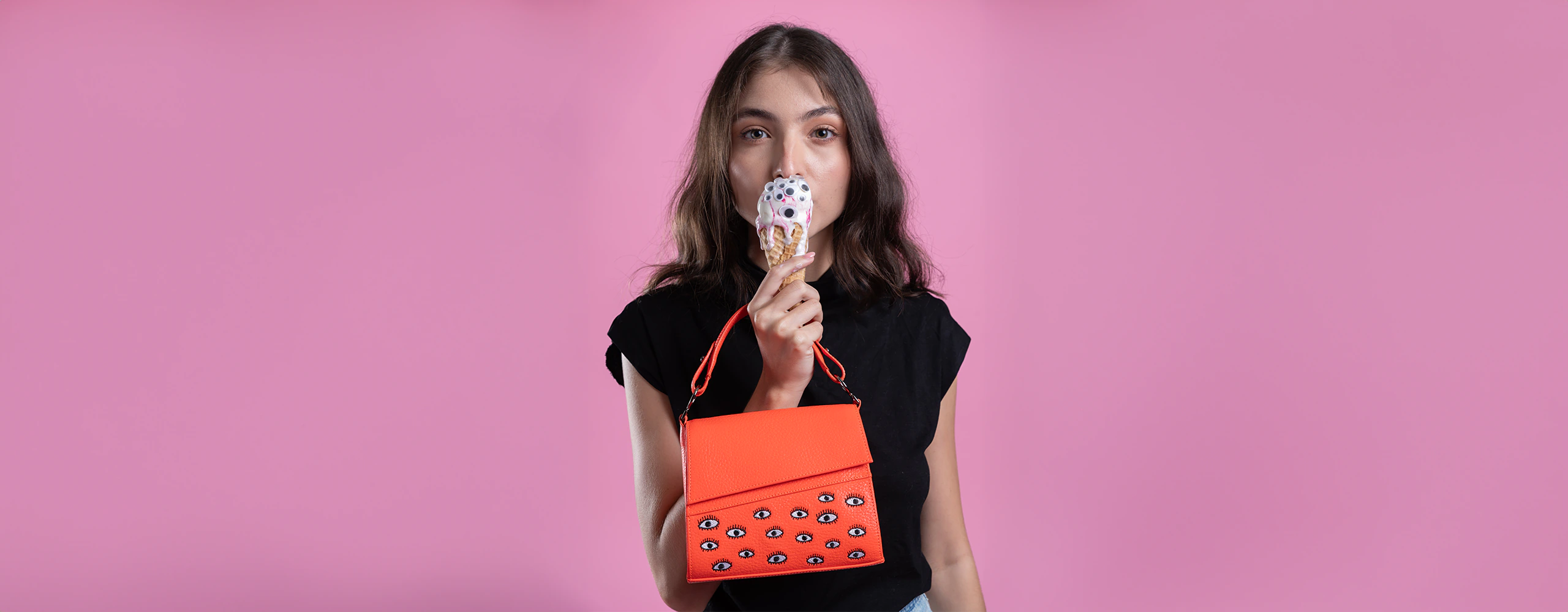 A young woman with light skin and long brown hair is holding an ice cream cone with googly eyes against a pink background. She is wearing a black top and holding an "Anastasio Crossbody Handbag" by Min & Mon, an NYC-based brand. The handbag is bright orange with a pattern of eye designs. The woman is looking directly at the camera while enjoying her ice cream.