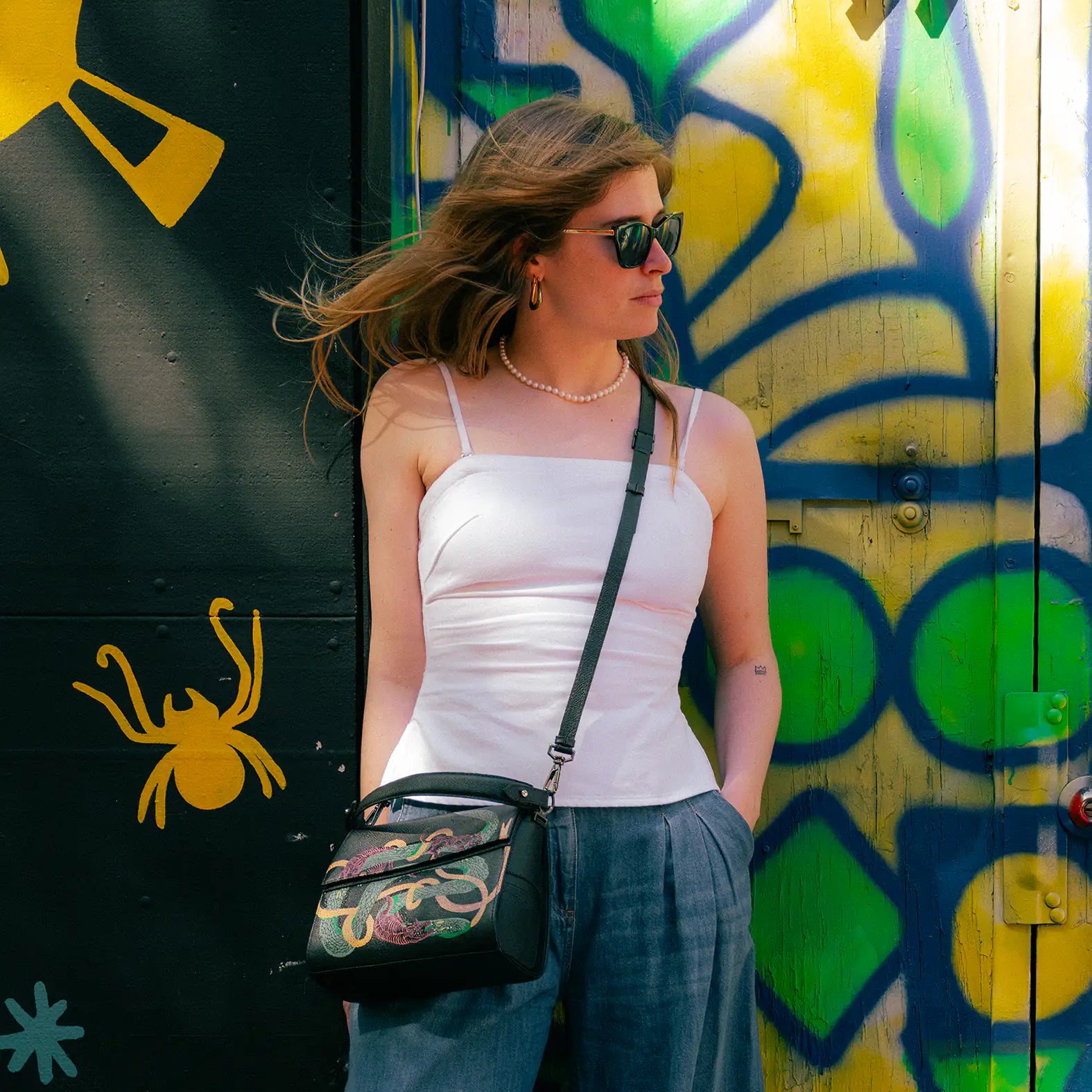 Woman in a white camisole and loose blue pants stands against a colorful graffiti wall, wearing sunglasses and a black crossbody bag with tigers and snake print design.