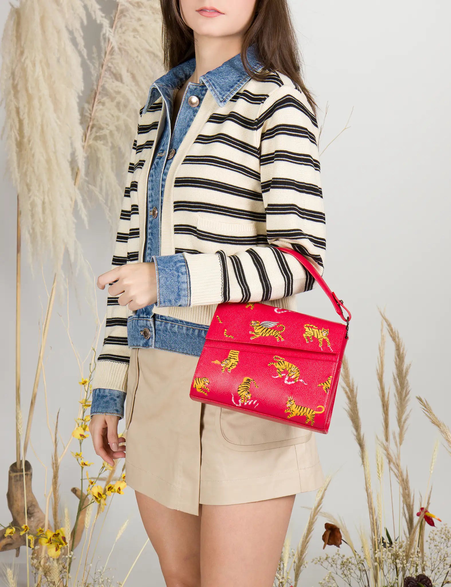 Woman wearing striped cardigan and beige shorts holds a red handbag with playful yellow tiger prints, styled with a top handle, against a backdrop of pampas grass and wildflowers.