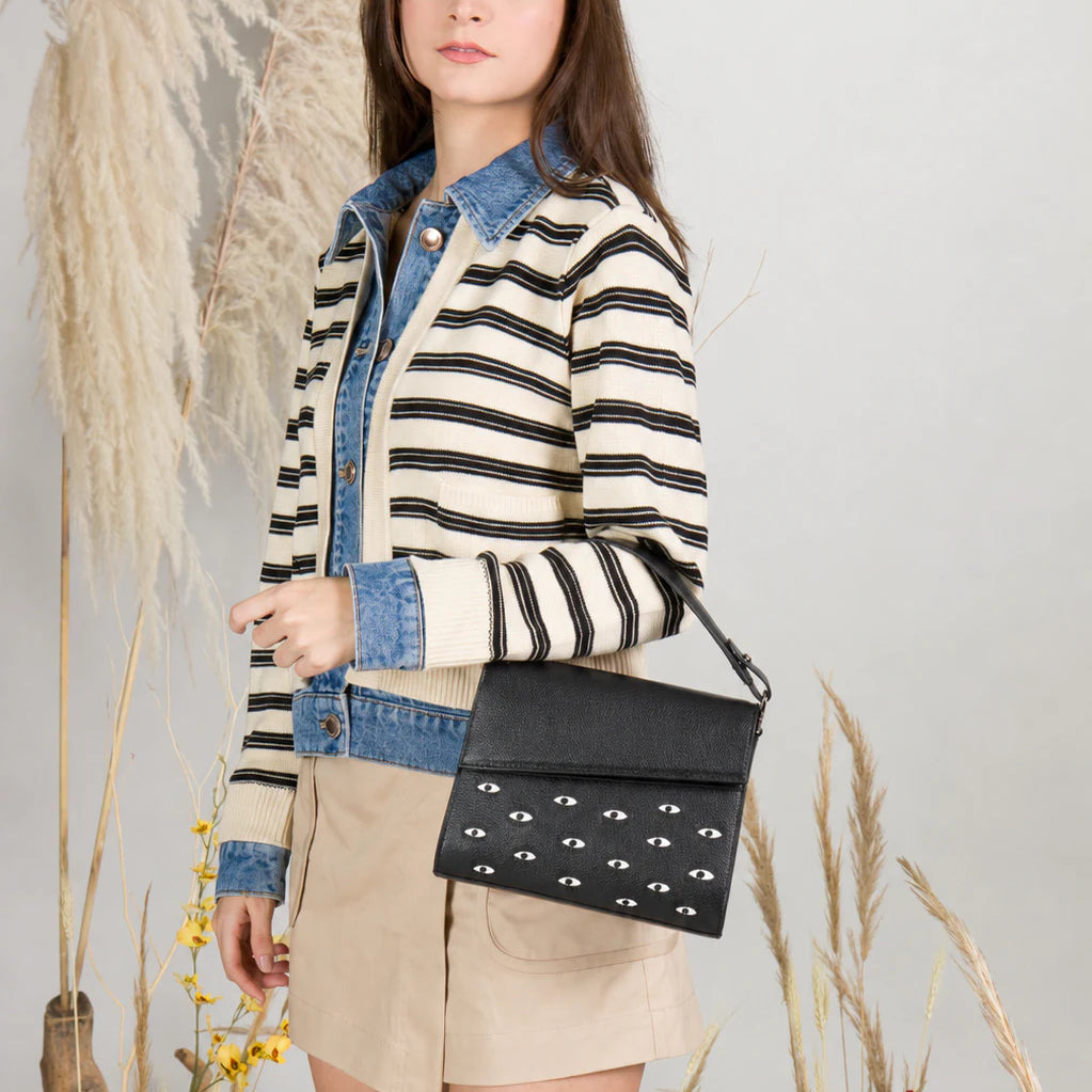Young woman in a striped jacket and beige skirt poses in a botanical studio, wearing a black shoulder bag with white eye motifs.