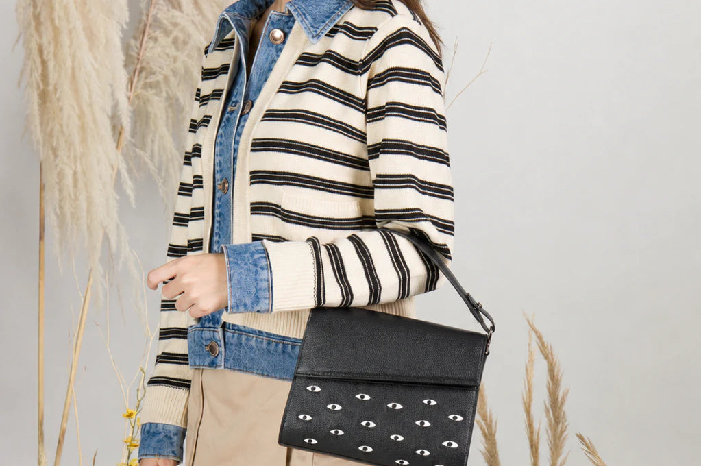 Young woman in a striped jacket and beige skirt poses in a botanical studio, wearing a black shoulder bag with white eye motifs.