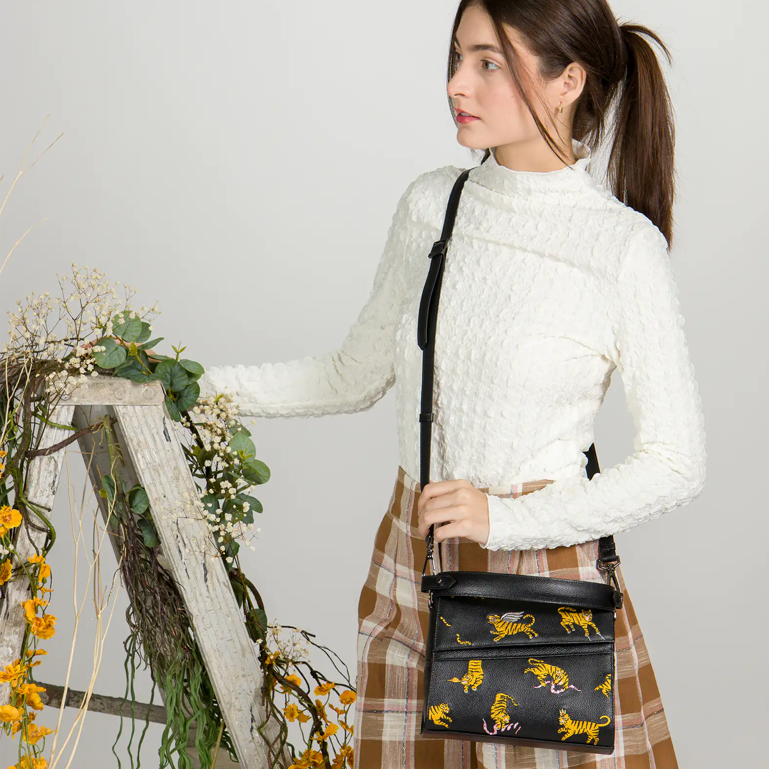 Woman in a white textured top and plaid skirt poses beside a rustic wooden ladder decorated with flowers, wearing a black crossbody bag with yellow tiger and pink snake embroidery.