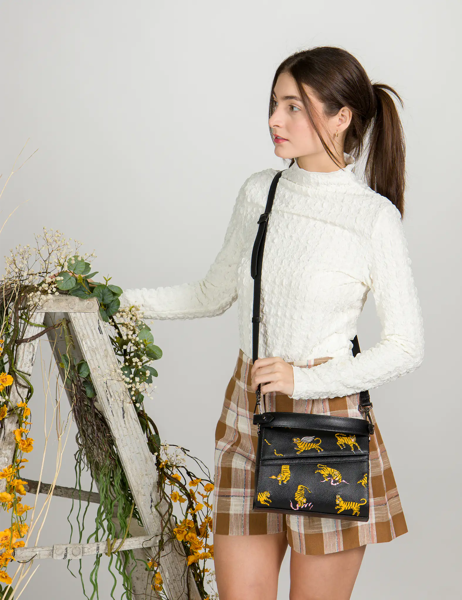 Woman in a white textured top and plaid skirt poses beside a rustic wooden ladder decorated with flowers, wearing a black crossbody bag with yellow tiger and pink snake embroidery.
