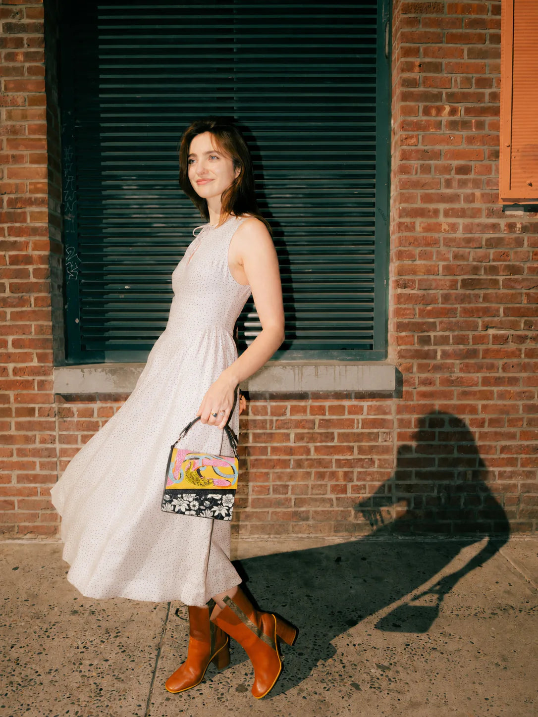 Woman in a flowing cream dress and tan heeled boots stands in warm sunlight against a brick wall, holding a colorful patterned handbag. A dramatic shadow stretches across the sidewalk behind her.