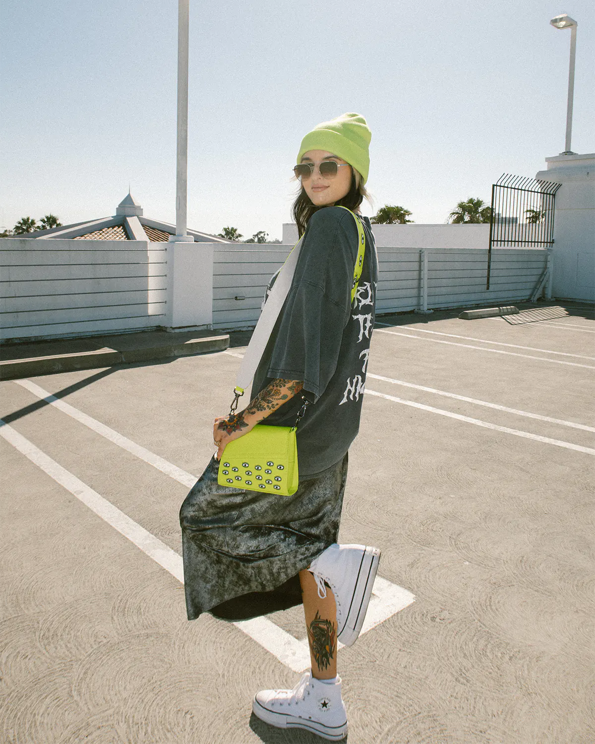 A stylish woman, sporting sunglasses and a neon yellow beanie, carries a matching neon Anastasio Micro Bag with eye motifs, blending effortlessly with her casual oversized graphic tee in a sunny rooftop setting.