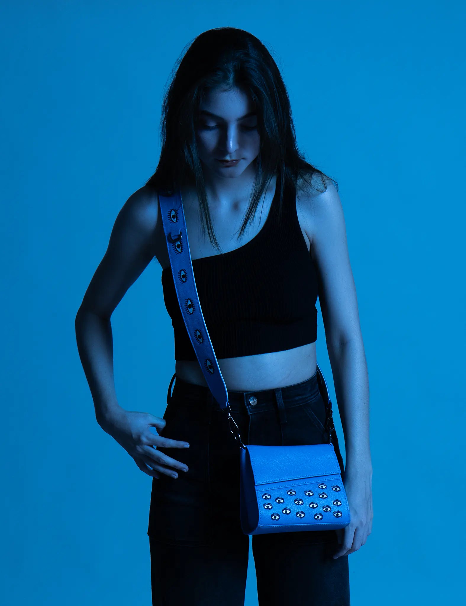 Model in a black crop top and dark jeans under blue lighting wears a crossbody bright blue mini handbag with eye embellishments and a wide strap, looking down.