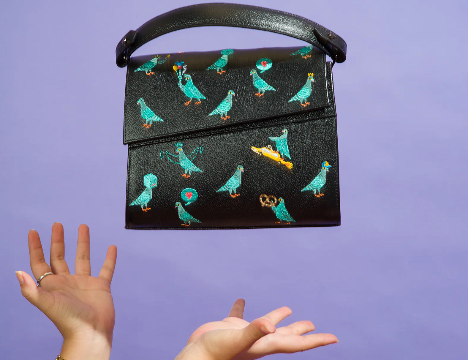 Tattooed arms toss a black handbag embroidered with playful teal pigeons, a pretzel, and a taxi, against a purple background. The bag floats mid-air with a curved top handle.