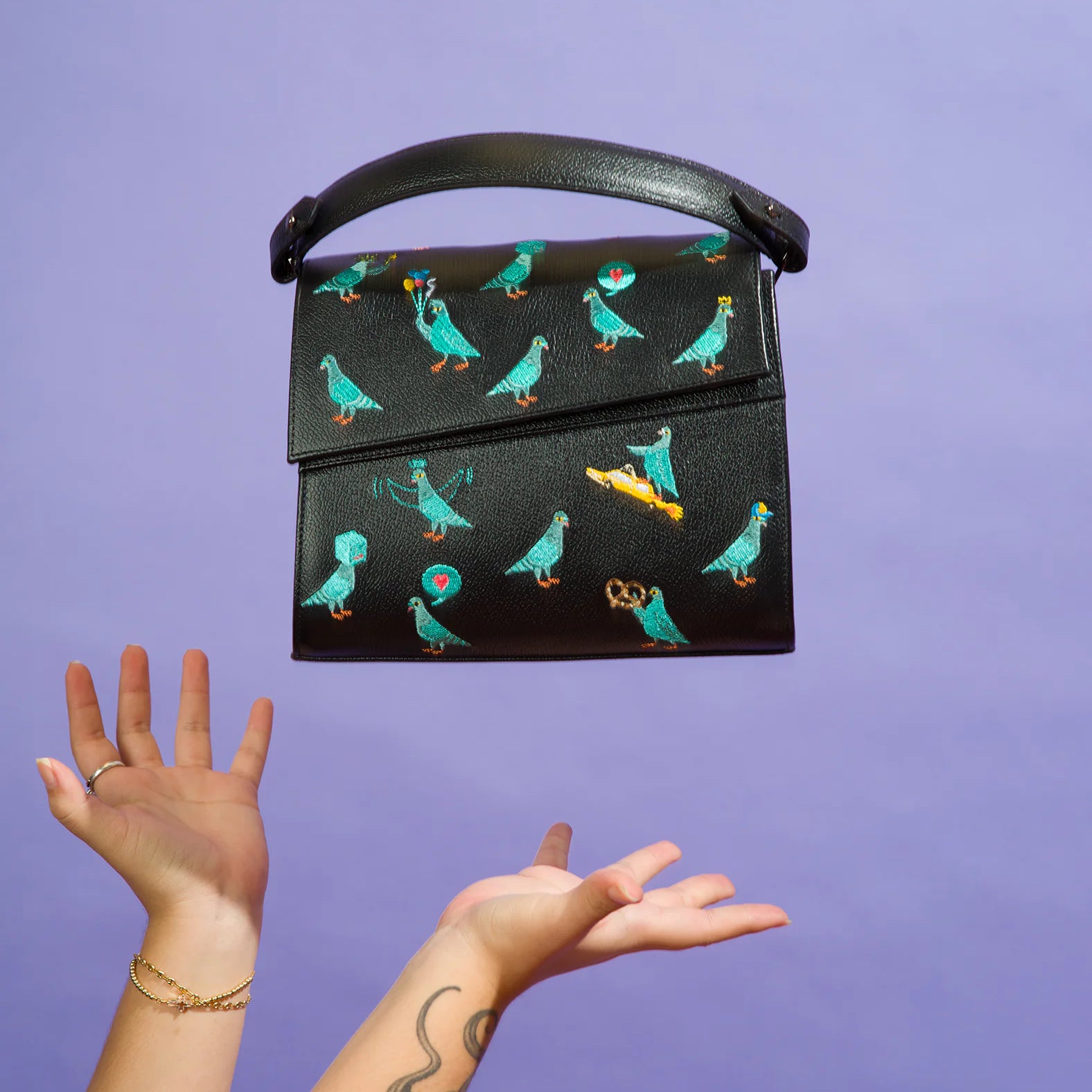 Tattooed arms toss a black handbag embroidered with playful teal pigeons, a pretzel, and a taxi, against a purple background. The bag floats mid-air with a curved top handle.