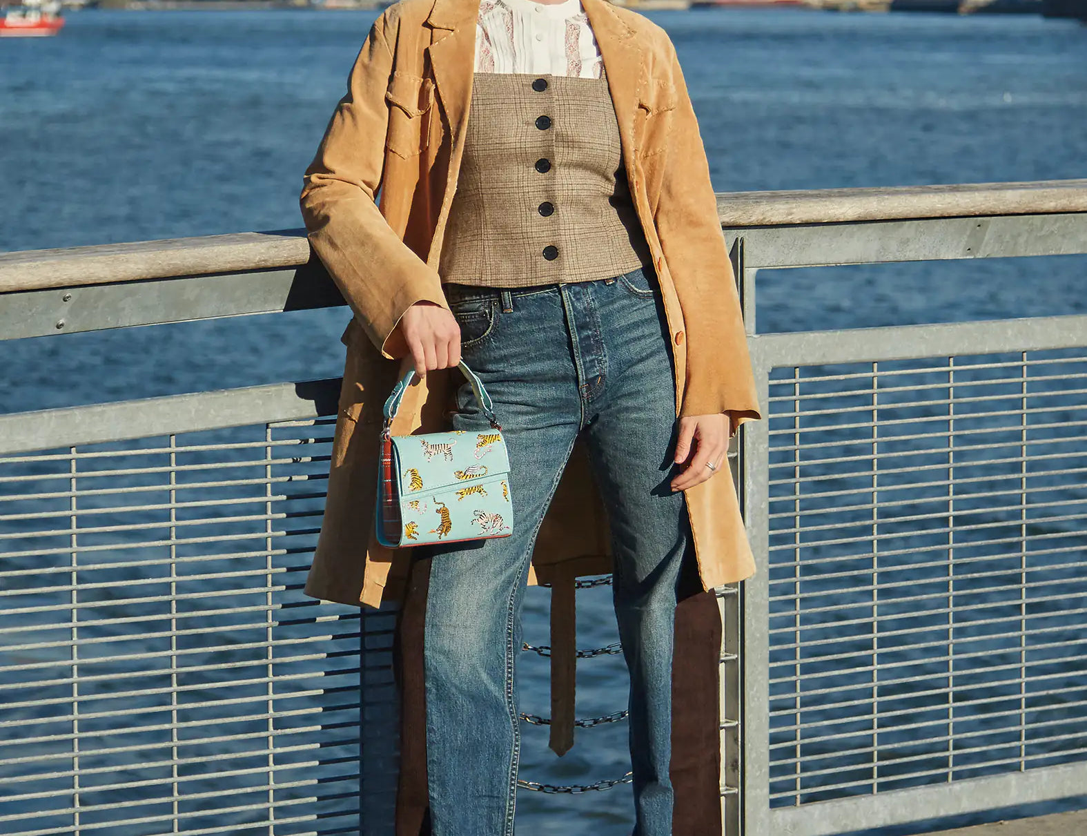 Woman standing by the waterfront wearing a tan coat, corset-style vest, and jeans, holding a light blue handbag embroidered with tigers.