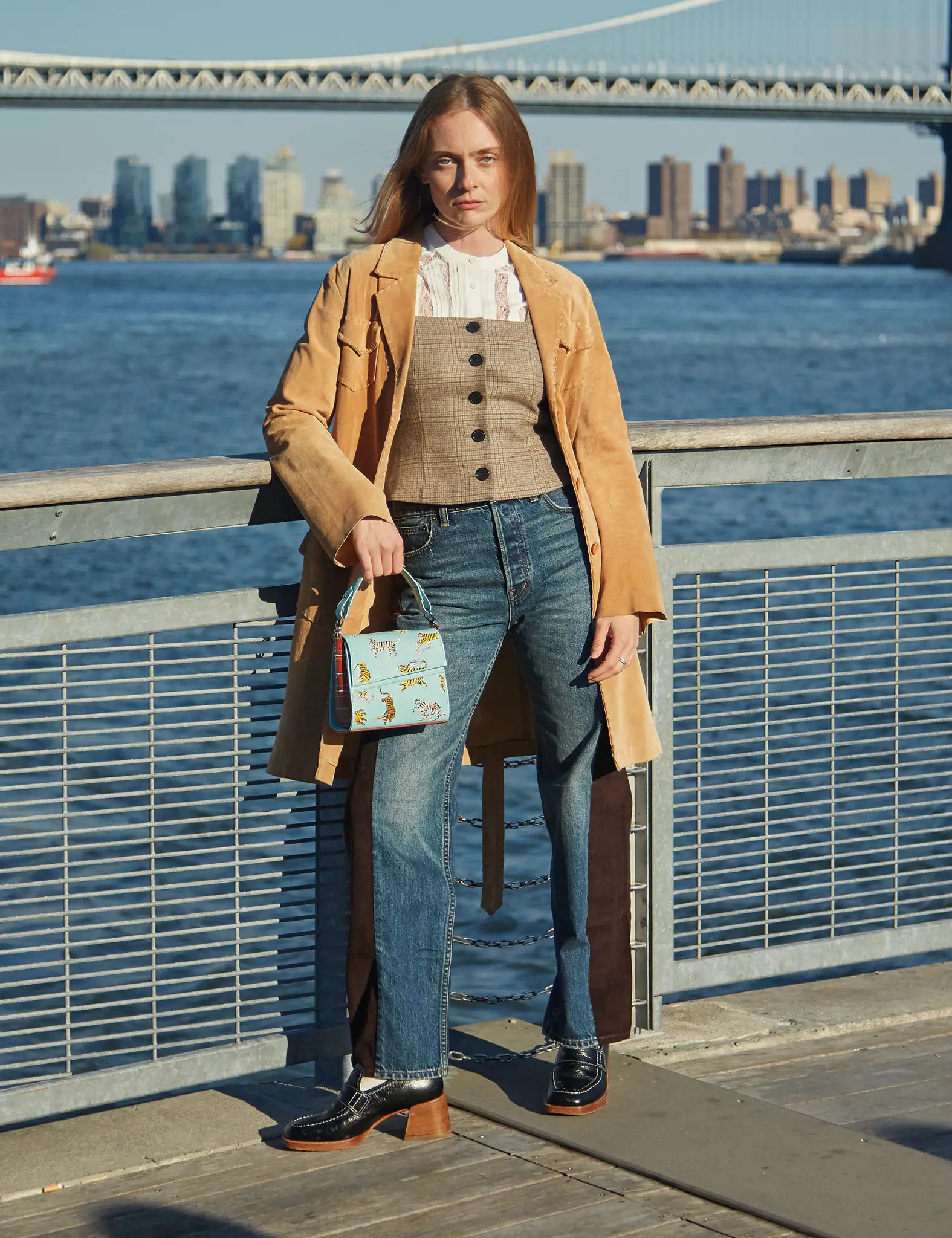 Woman standing by the waterfront wearing a tan coat, corset-style vest, and jeans, holding a light blue handbag embroidered with tigers.