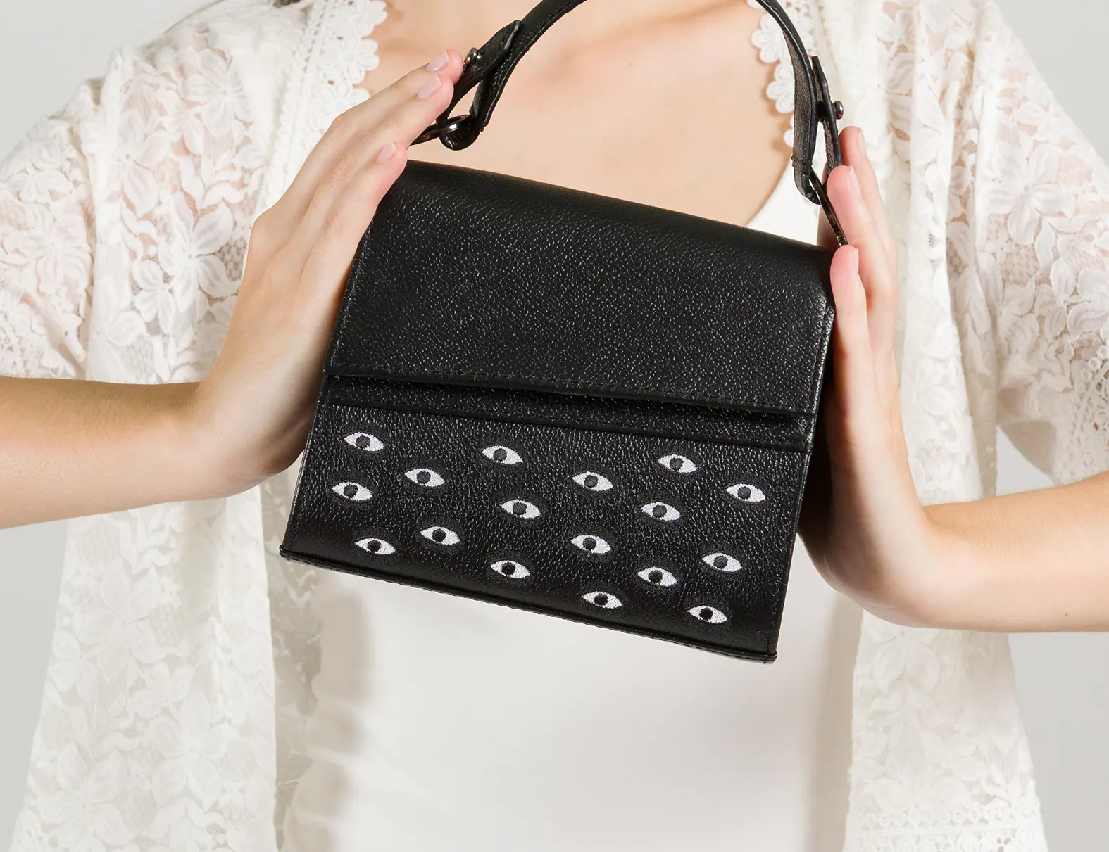 Model in a white dress and lace cardigan holding a structured black handbag adorned with a pattern of small white eyes, against a plain light background.