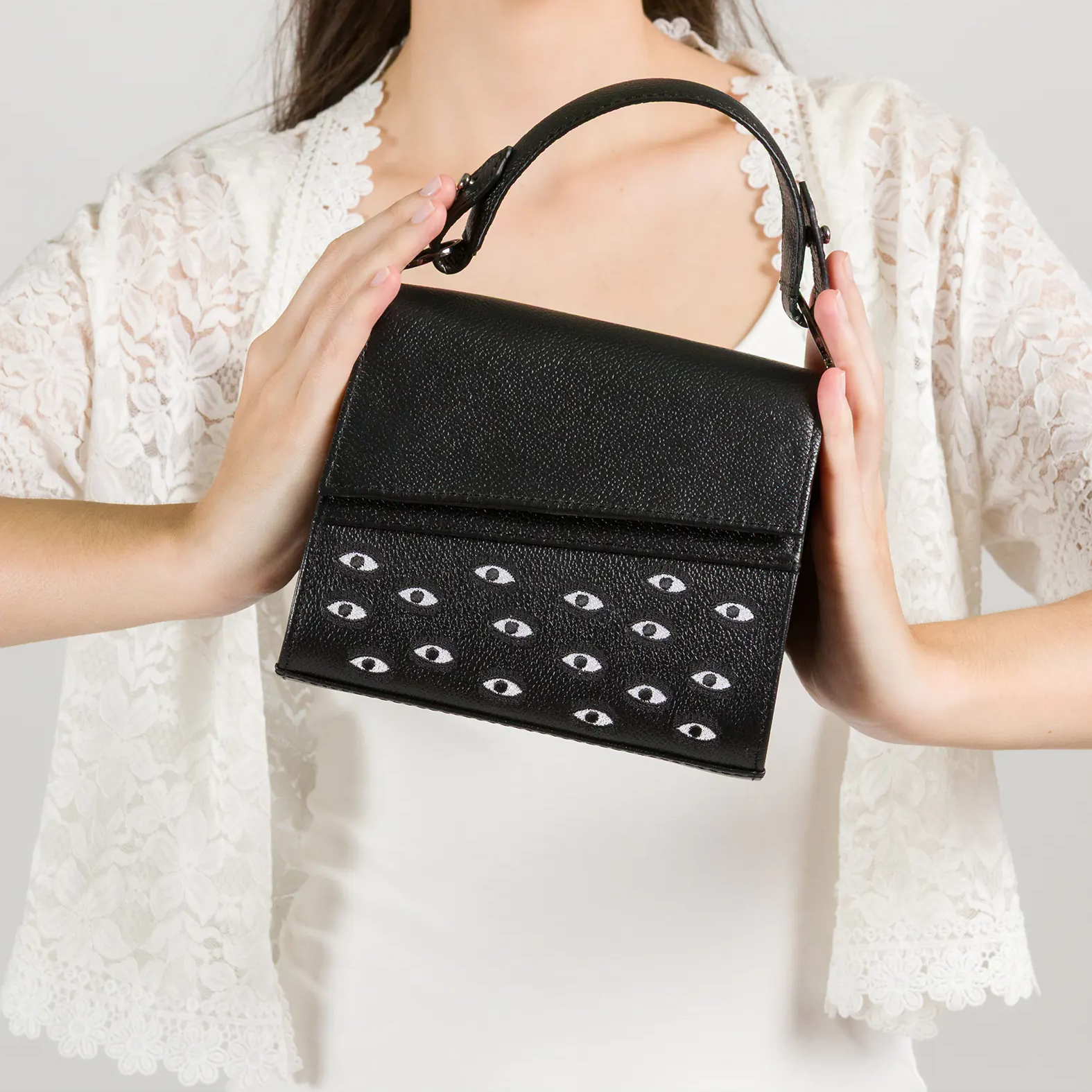 Model in a white dress and lace cardigan holding a structured black handbag adorned with a pattern of small white eyes, against a plain light background.