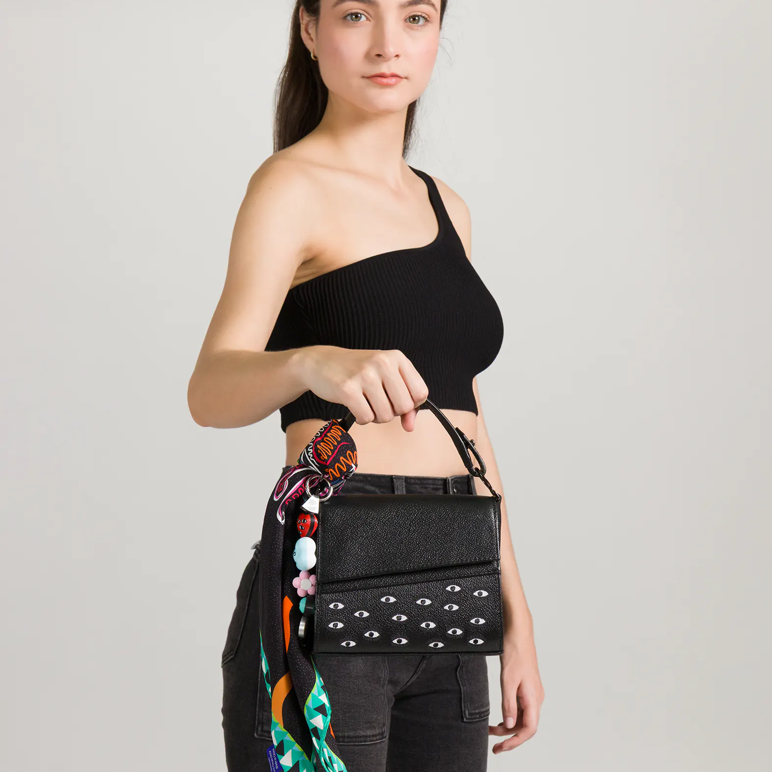 Model in a black one-shoulder crop top and dark jeans holding a black handbag with white eye patterns, decorated with a colorful scarf and charms, against a plain light background.