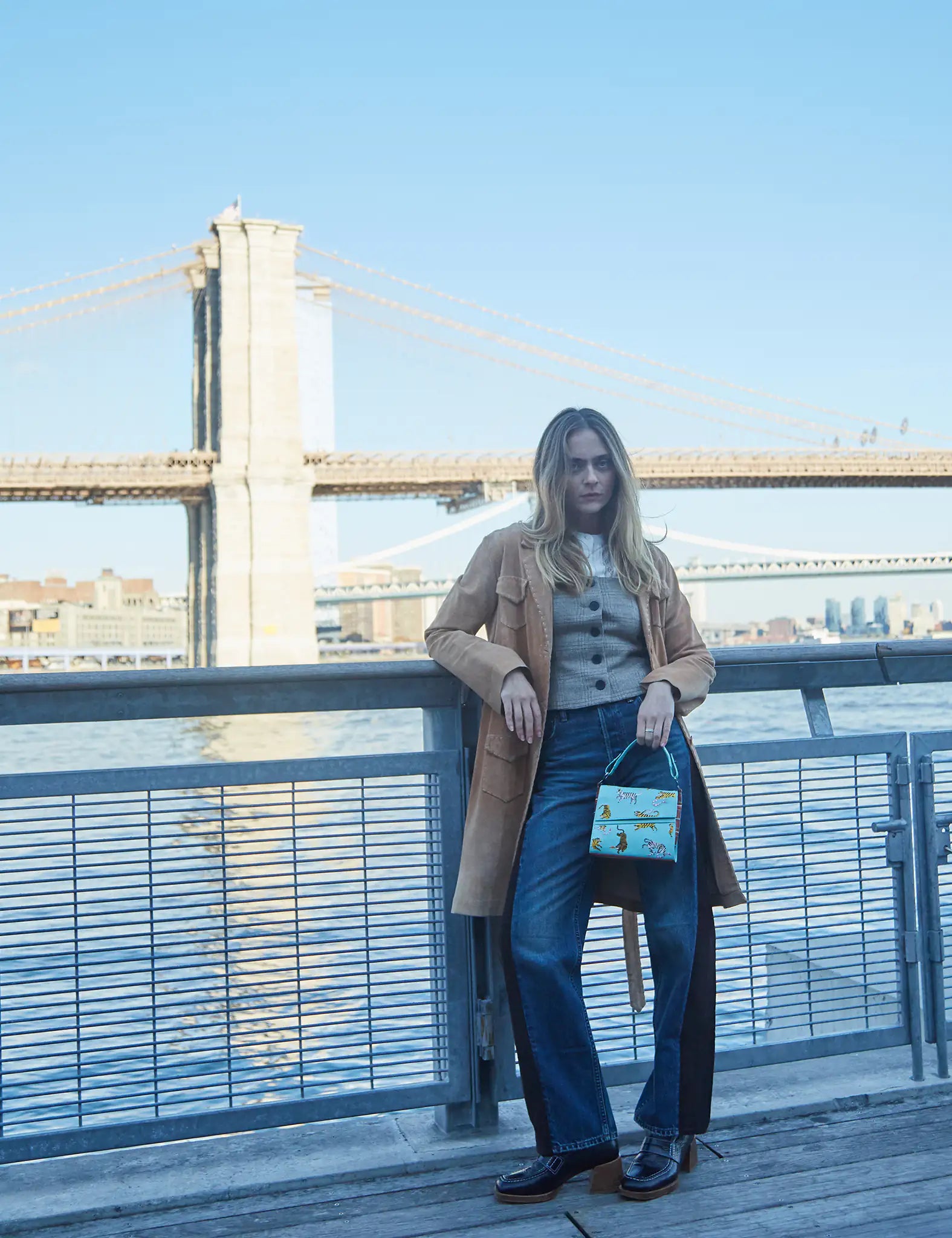 Woman standing by a river railing near a bridge, wearing a tan coat and jeans, holding a light blue handbag embroidered with yellow tigers and pink snakes.