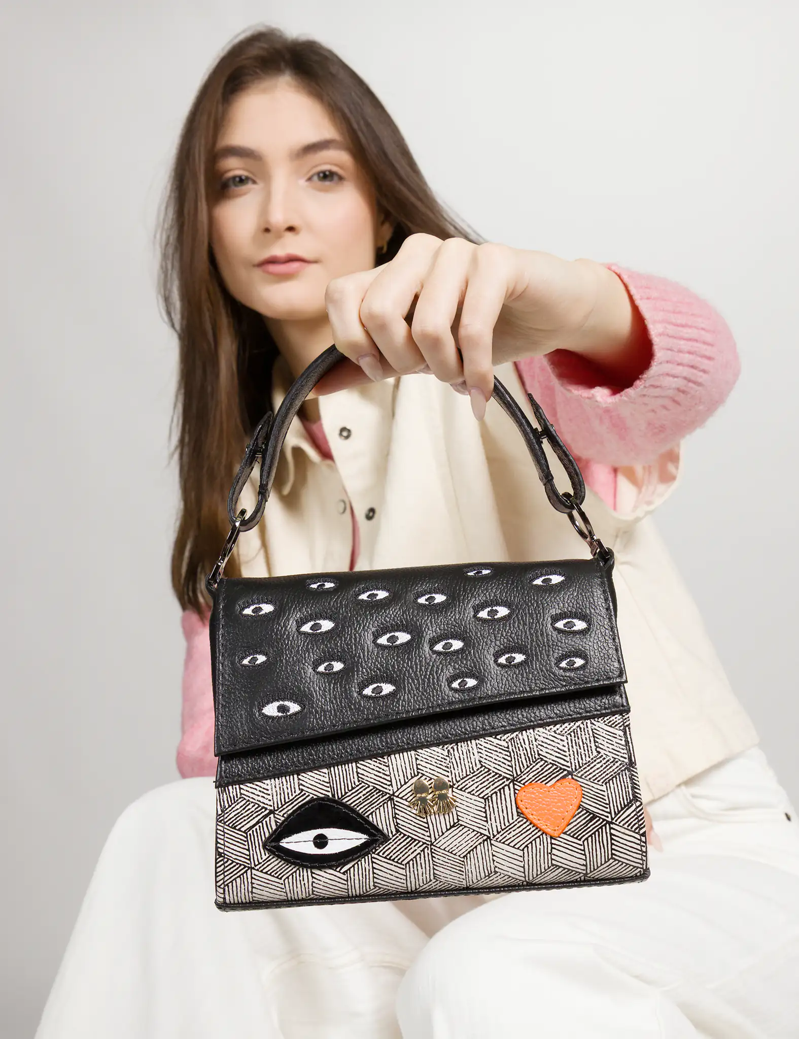 A woman holds a structured handbag toward the camera. The bag features a black flap embroidered with multiple small eyes, while the bottom half has a geometric beige-and-black pattern accented with an orange heart and a large eye motif. She wears a pink sweater layered with a cream vest and white pants.