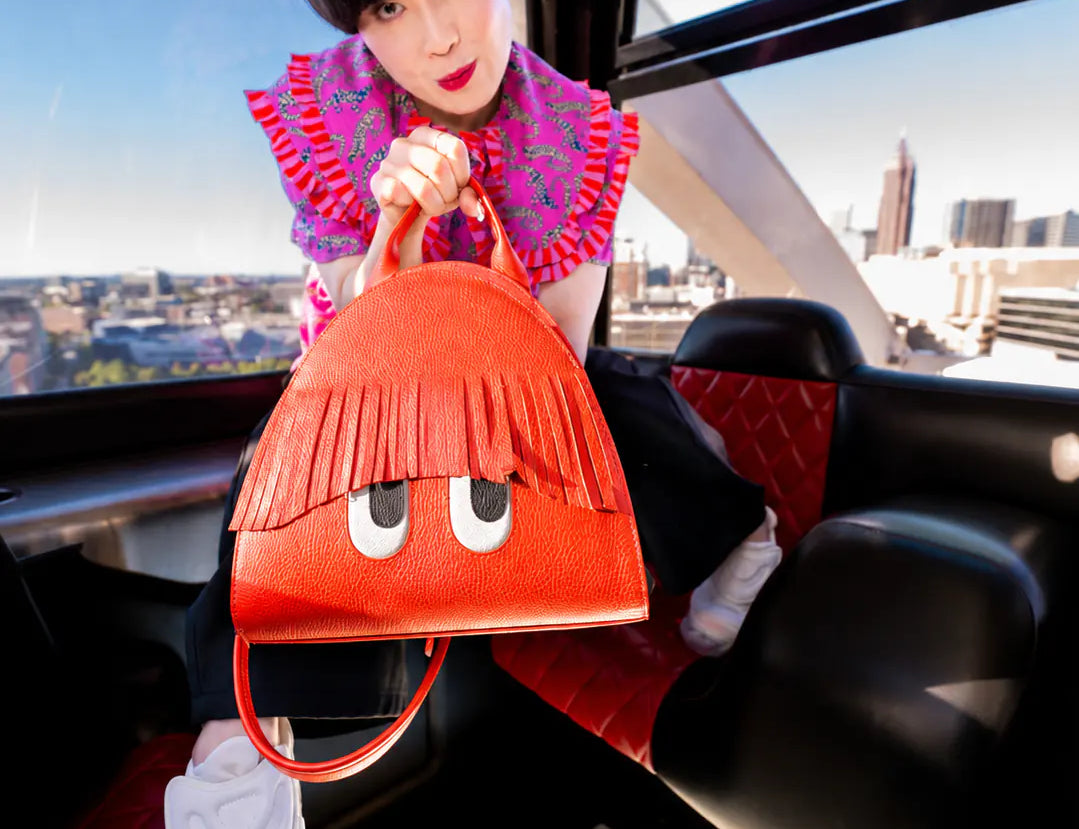 Model in a vibrant pink patterned top showcases an orange Min & Mon handbag with playful fringe bangs and engraved eyes while posing inside a ferris wheel car overlooking the city.